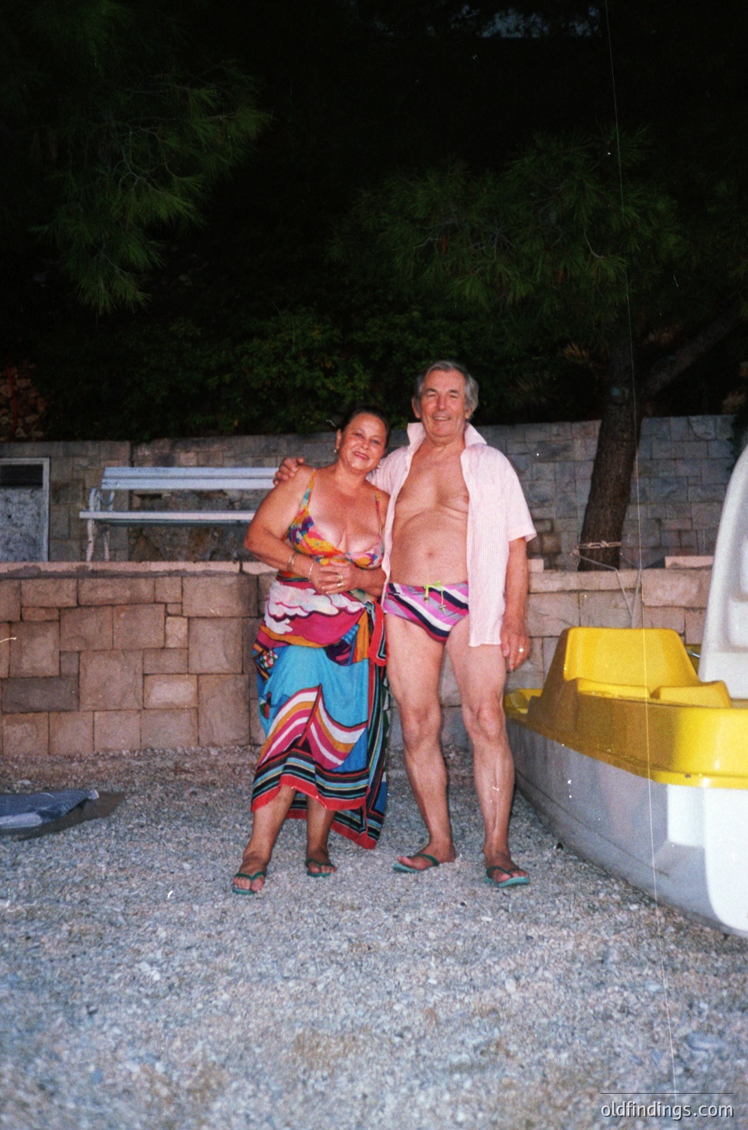 Two adults pose casually in a retro beach setting, likely Mediterranean. The woman wears a colorful, patterned swimsuit with a sarong-style skirt, while the man dons a pink robe over red swim trunks. A yellow portable toilet and stone wall with greenery suggest a seaside resort or private pool area. Style hints at 1970s–1980s beach culture.