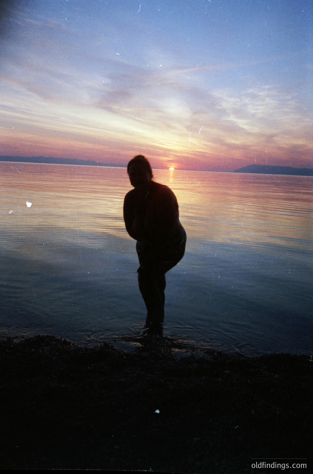 Silhouetted figure stands at water’s edge during golden-hour sunset, back to camera. Warm tones reflect on calm waters, likely a lake or coastal inlet. Vintage film grain suggests mid-20th century (1950s–1970s).