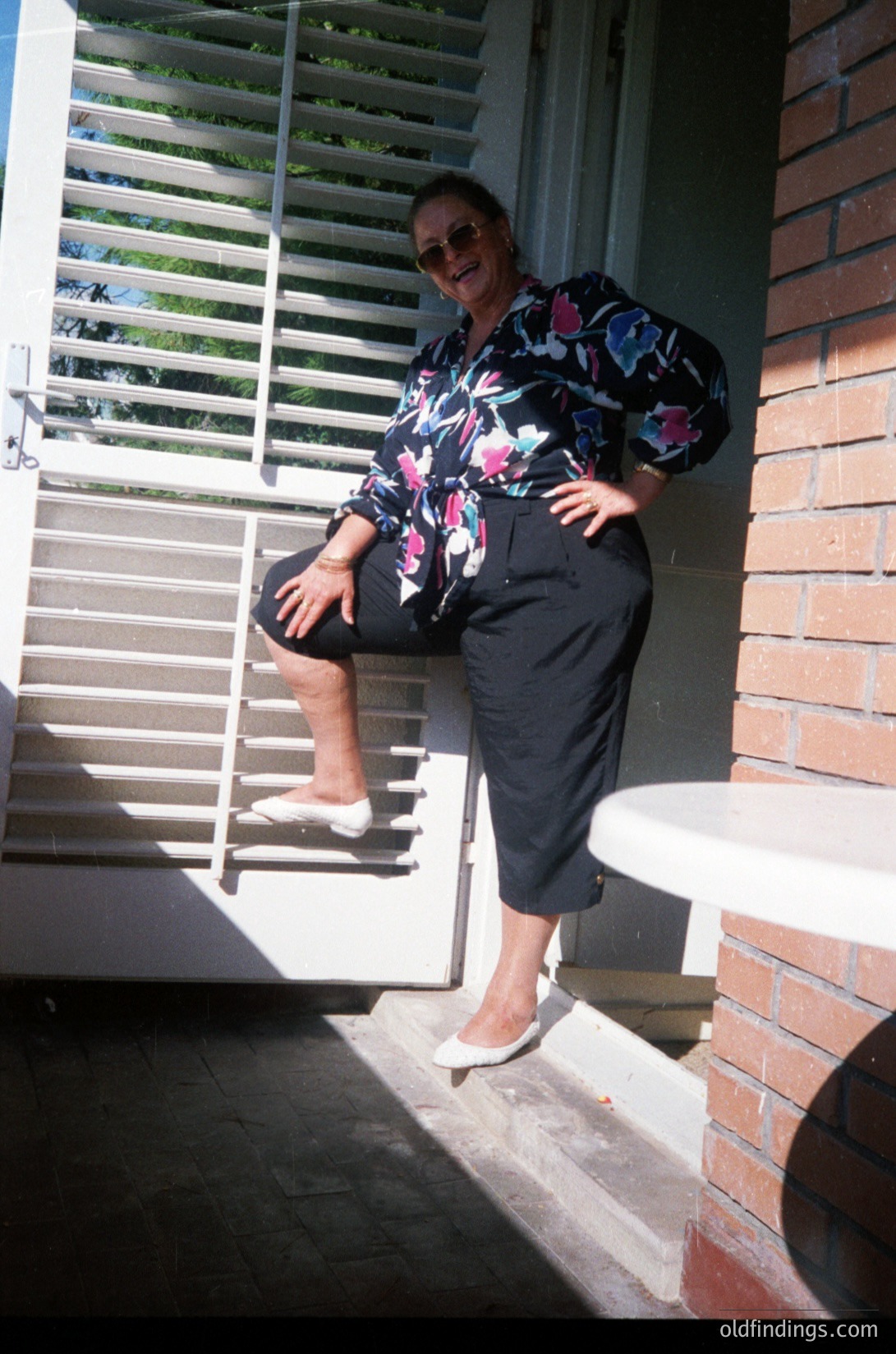 Vintage 1970s portrait of a woman posing on brick steps beside a sliding glass door with louvered blinds. She wears a floral-patterned blouse, high-waisted wide-leg trousers, white sandals, and sunglasses. The setting suggests suburban residential architecture.