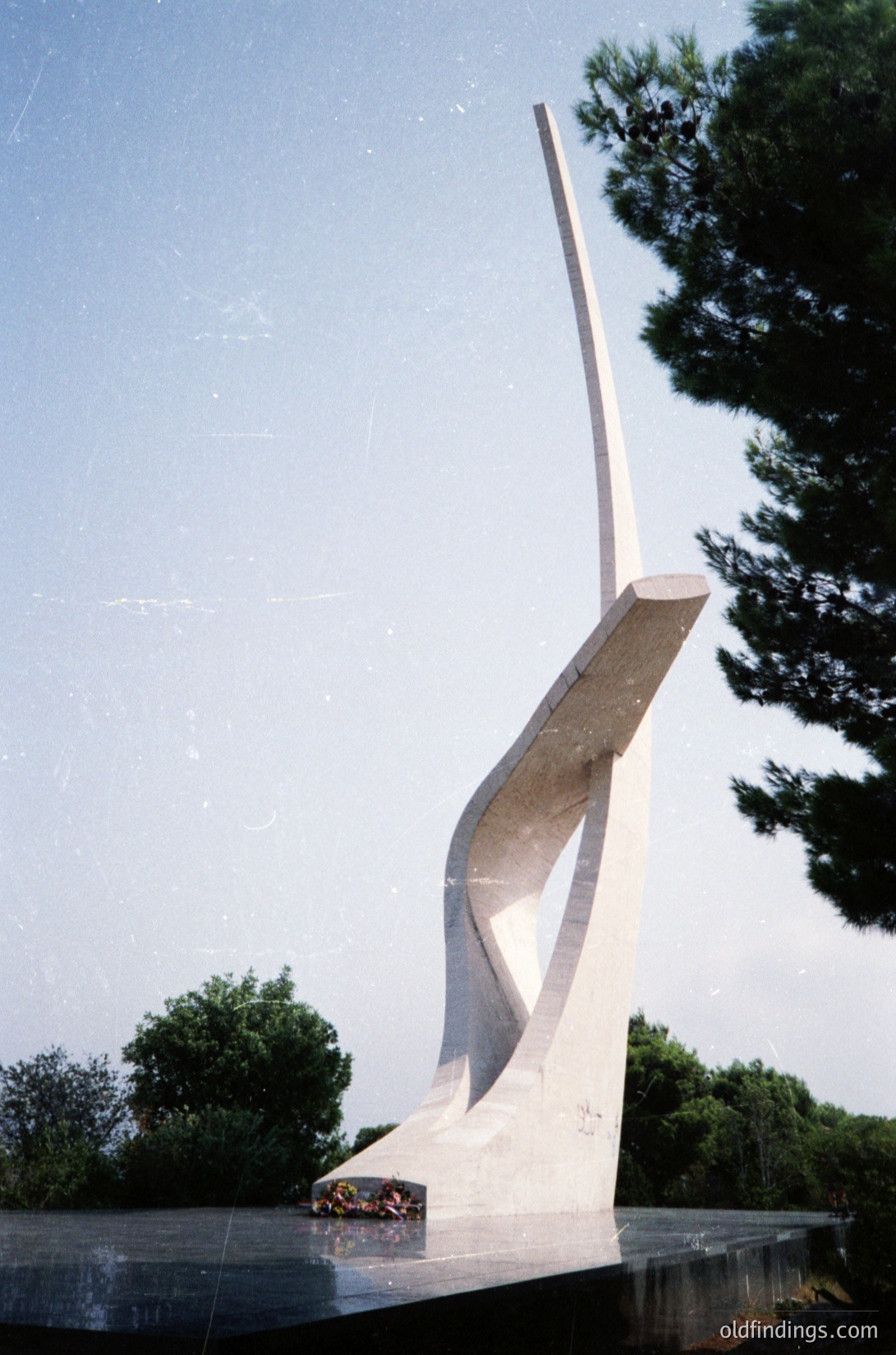 Modernist concrete monument resembling a stylized flame or abstract wing, set on a circular base. Minimalist design with clean lines and smooth surfaces. Surrounded by greenery and a clear sky, suggesting an outdoor public space.