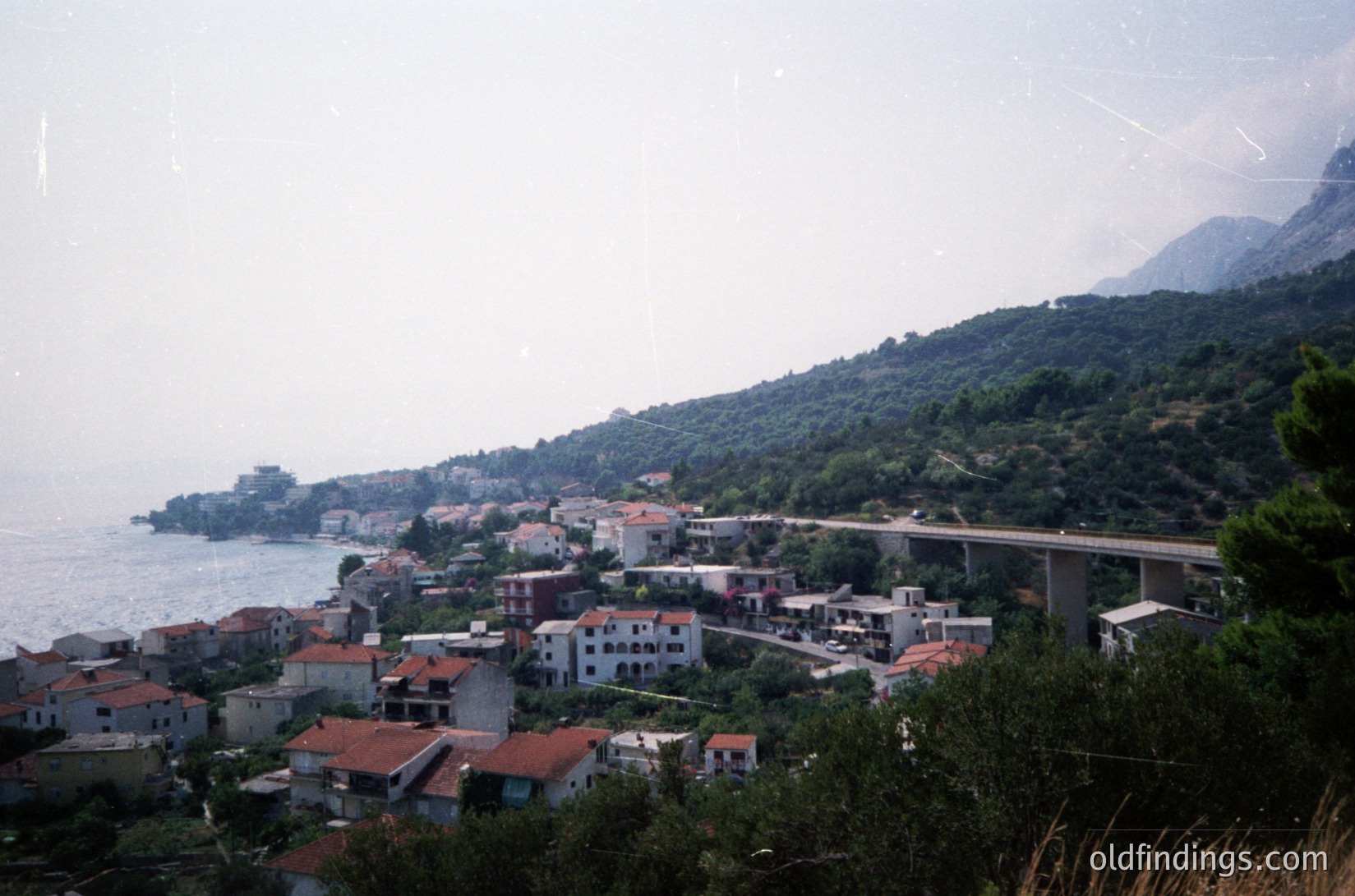 Coastal village nestled between sea and mountains, featuring clustered Mediterranean-style buildings with terracotta roofs. A curved road bridge spans the coastline, connecting settlements. Lush greenery and rocky terrain frame the scene, suggesting a rugged Mediterranean coastline. Likely –1980s based on architectural style and film grain.