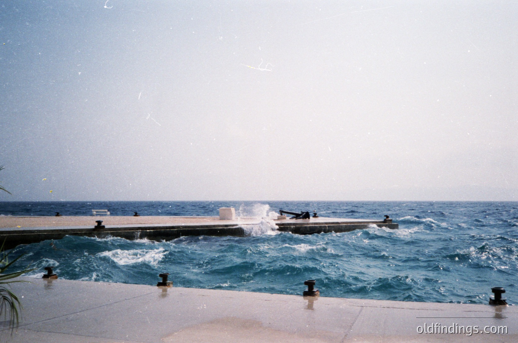Vintage seaside pool with concrete edge and wooden deck extending into choppy waves. Two figures in swimwear stand near the water’s edge, while a third sits on the deck. Mid-20th century design with black metal railings and spherical bollards.