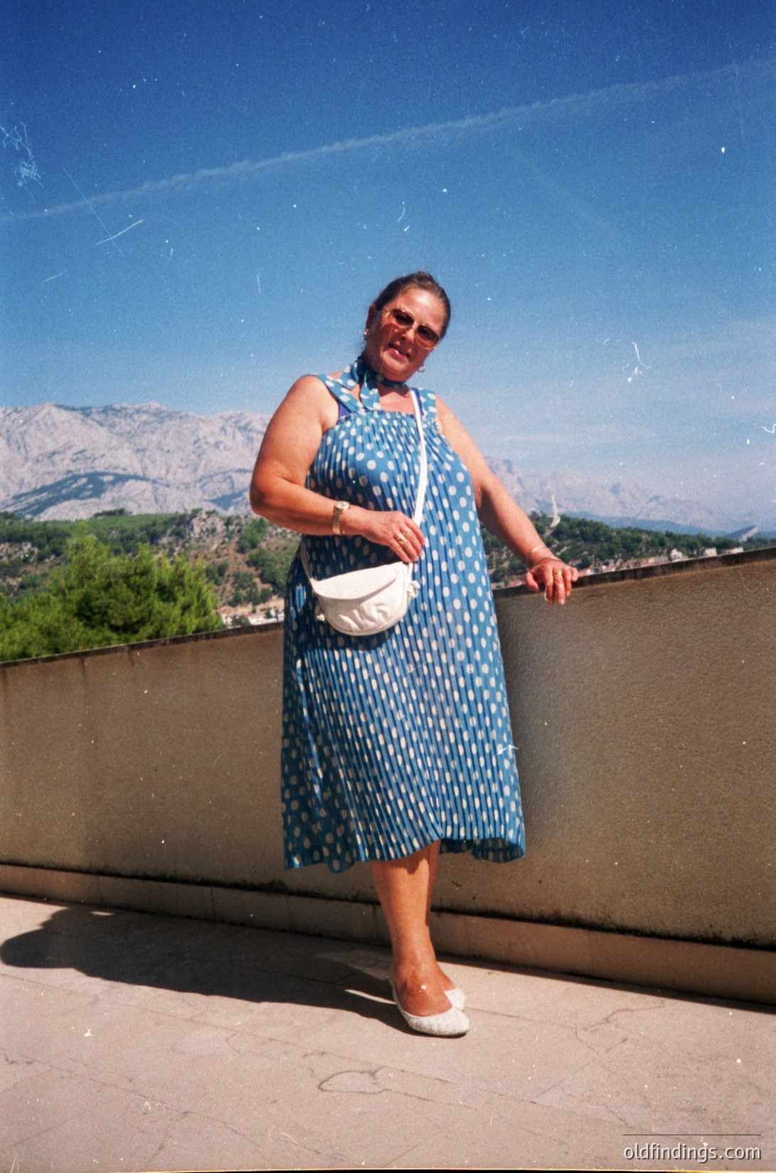 A woman in a 1990s-style polka-dot dress poses on a rooftop terrace with alpine mountains in the background. She wears a white crossbody bag and sandals, leaning against a concrete railing. The image captures a bright, sunny day, likely in a European alpine region.