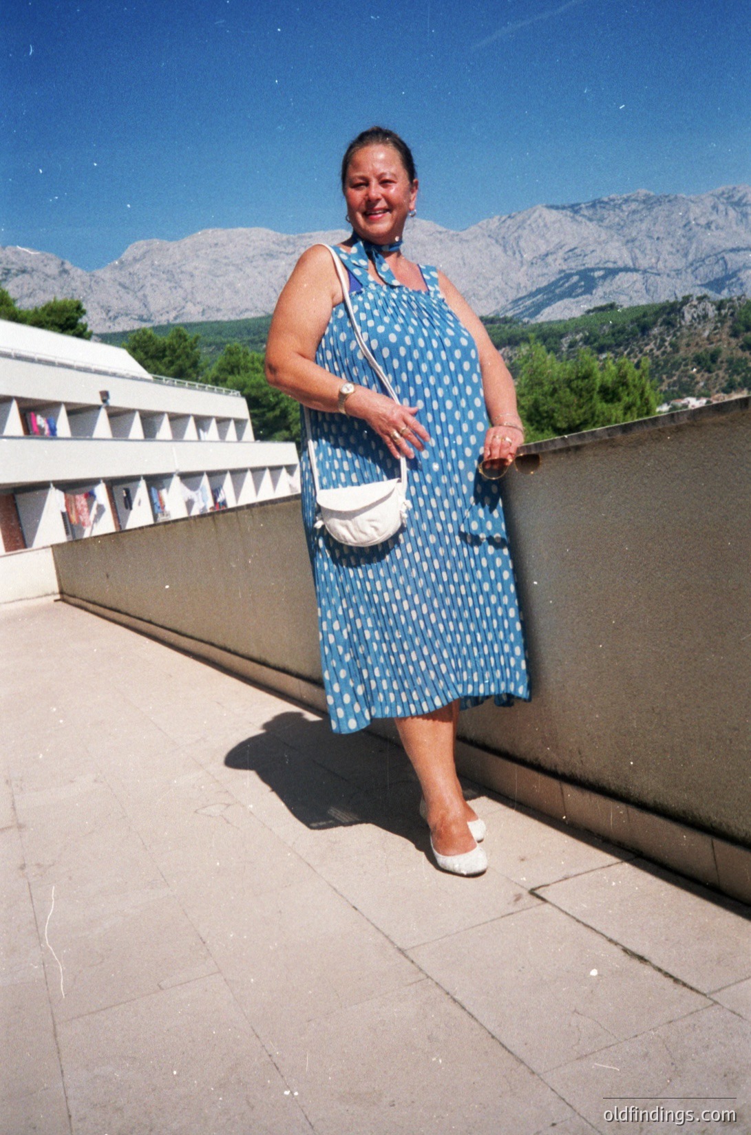 Woman in a 1970s-style blue polka-dot dress poses outdoors beside a concrete barrier, set against mountainous alpine scenery. She wears a white crossbody bag and sandals, with a watch on her wrist. Likely taken in a European alpine region.