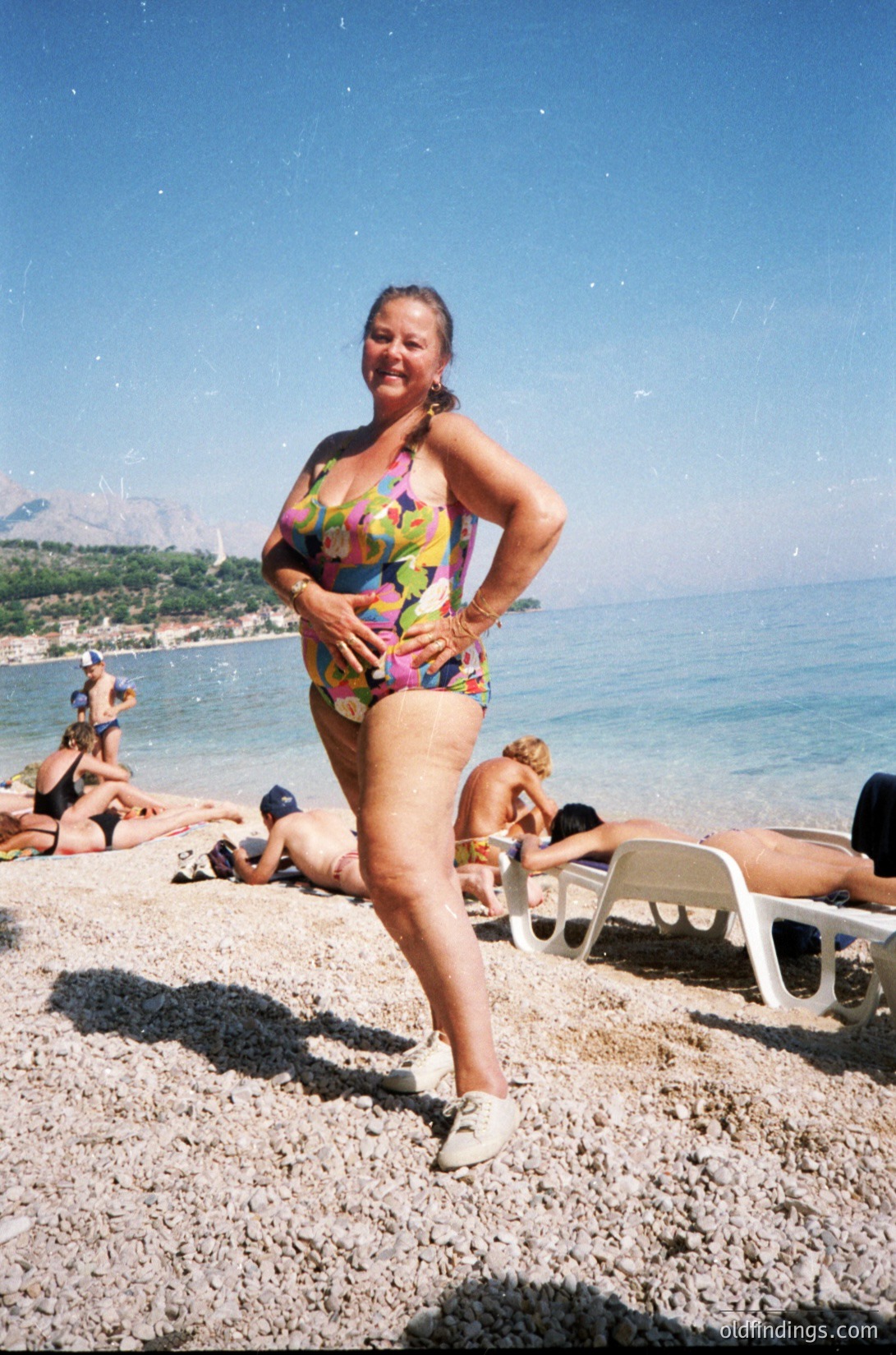 Vintage seaside scene featuring a woman in a colorful floral swimsuit posing on pebble beach. White plastic lounge chairs and sunbathers in background. Coastal mountains and clear blue sea in distance. Likely Mediterranean or Adriatic region, 1980s-1990s.
