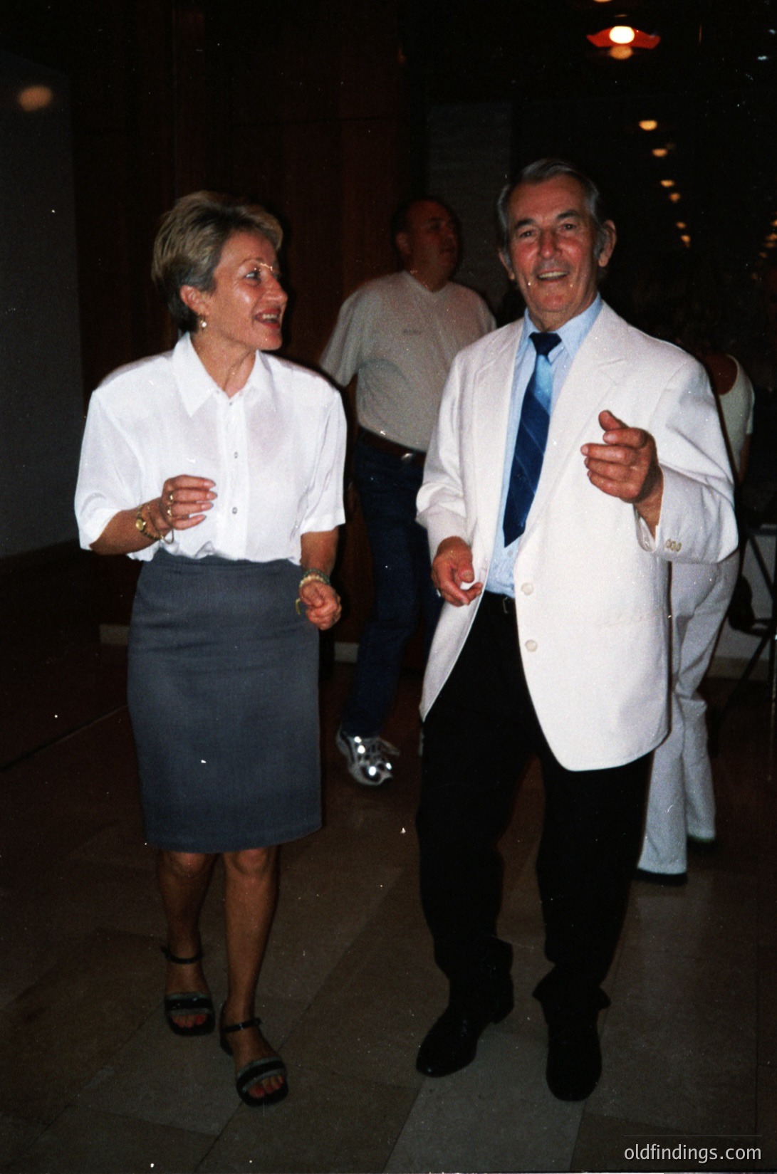 Two elegantly dressed adults laughing indoors at an evening event, likely 1990s–2000s. Woman in a white blouse, dark skirt, and sandals; man in a white blazer, dress shirt, and striped tie. Soft lighting, marble flooring, and blurred background suggest a formal reception or gala.