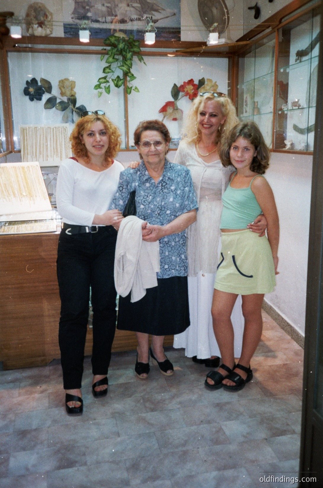 Four women pose indoors in a vintage clothing store, likely 1980s–1990s. The elder woman wears a patterned blouse and black skirt; the others display 80s fashion—cropped tops, high-waisted shorts, and sandals. Shelves display mannequins in dresses and floral arrangements. Warm lighting and retro decor suggest a European boutique.