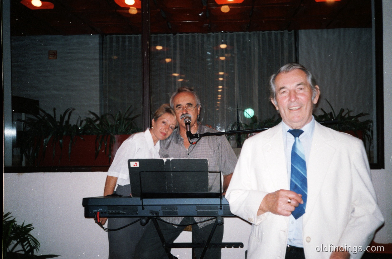 Indoor evening event featuring three performers on a stage with modernist decor. A man in a white suit and blue tie stands front-left, gesturing toward the audience. Behind him, a woman in a white blouse plays a keyboard, while a man in a gray shirt sings into a microphone. Potted plants and sheer curtains frame the background, suggesting a mid-20th century venue.