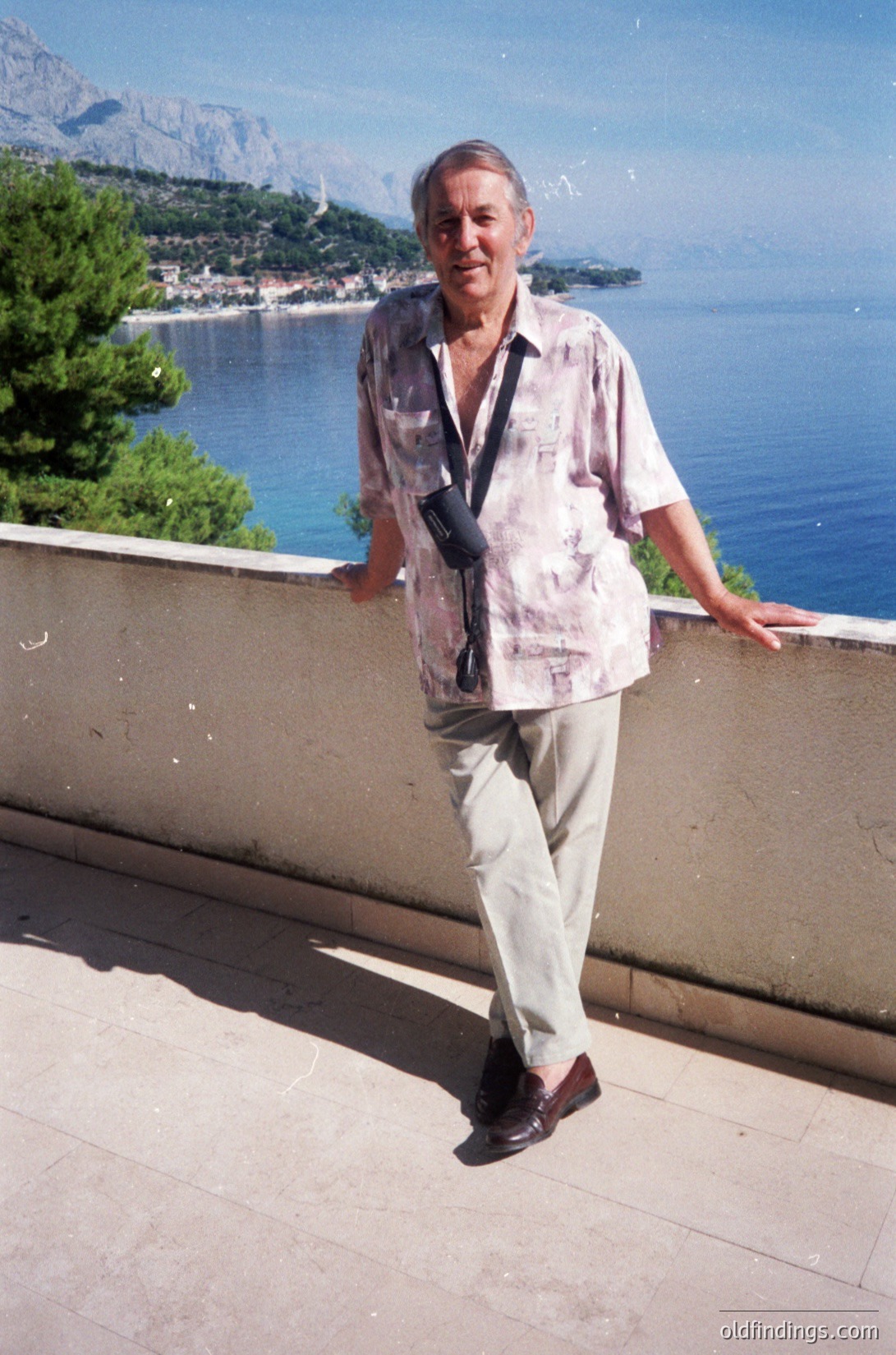 Mid-20th century man poses on a sunlit terrace overlooking a coastal town and deep blue sea, framed by lush greenery and rocky mountains. Light-colored linen shirt, beige trousers, and dark shoes suggest a Mediterranean or European seaside locale, likely the 1960s–1980s. Camera strap indicates travel or photography hobby.