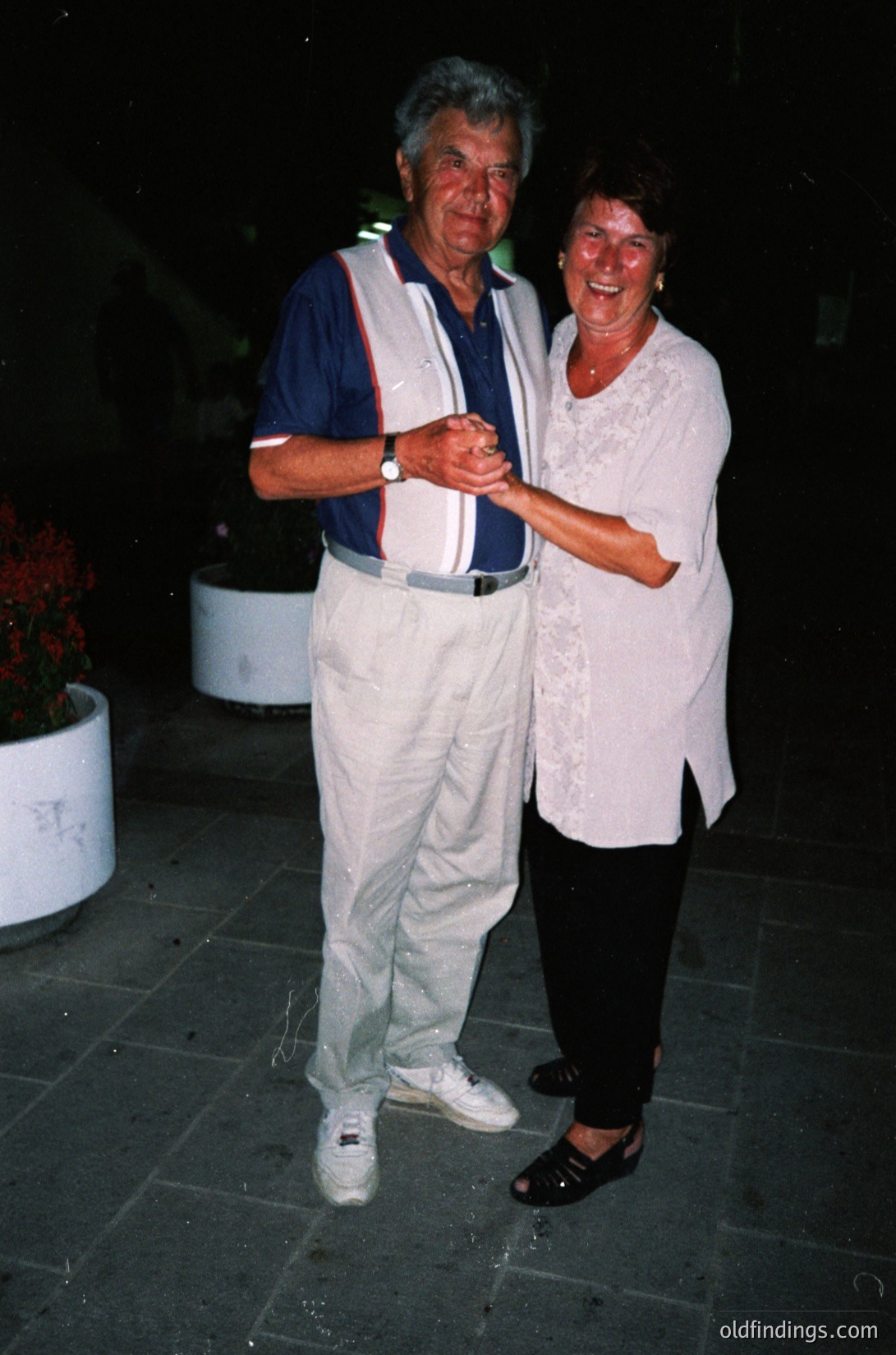 A man and woman pose closely outdoors at night, likely in the 1980s–1990s. The man wears a striped athletic vest, white pants, and a watch; the woman wears a white lace top and black pants. They stand on a tiled patio with potted plants and blurred greenery in the background. [Retro couple posing ]