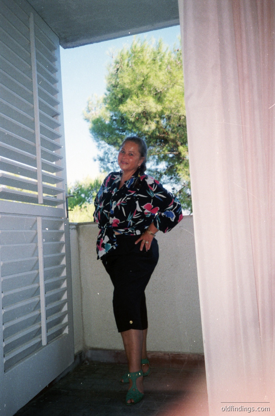 Woman in floral-patterned blouse and high-waisted skirt poses in a sunlit hallway, framed by white louvered doors. Green sandals and a relaxed stance suggest mid-20th-century fashion. Lush greenery outside hints at a Mediterranean or coastal setting.