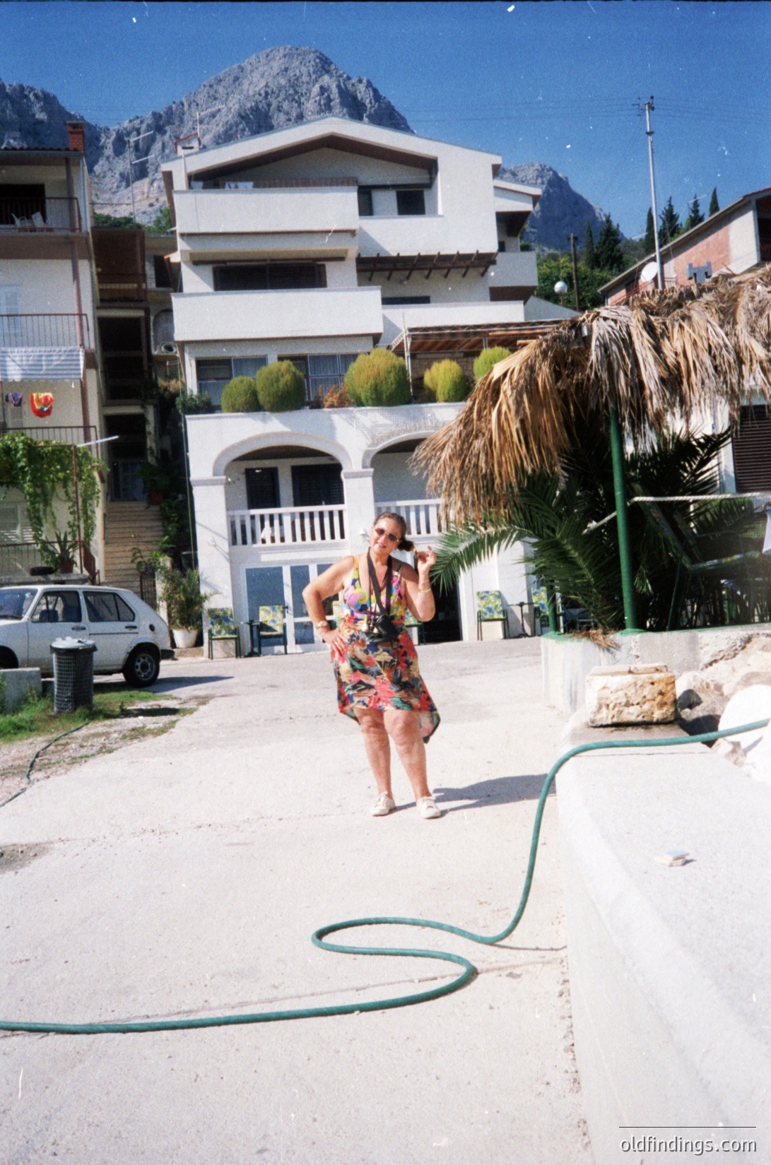 Woman in a floral dress uses a green hose on a paved driveway beside a two-story residential building with arched windows, set against mountainous terrain. Tropical plants and a thatched awning frame the scene. Likely a seaside or alpine resort area, 1990s–2000s.