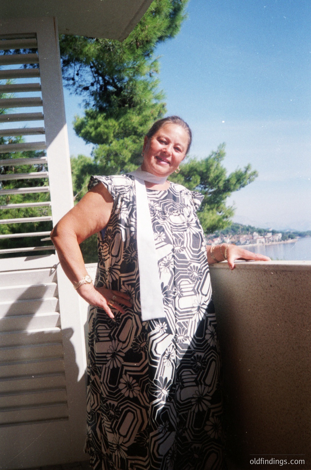 Woman in a patterned, knee-length dress with geometric motifs poses on a balcony overlooking a waterfront, likely a coastal town. The dress features a white collar and sleeves, suggesting a 1970s–1980s style. Lush greenery and a clear sky frame the scene, indicating a sunny day.