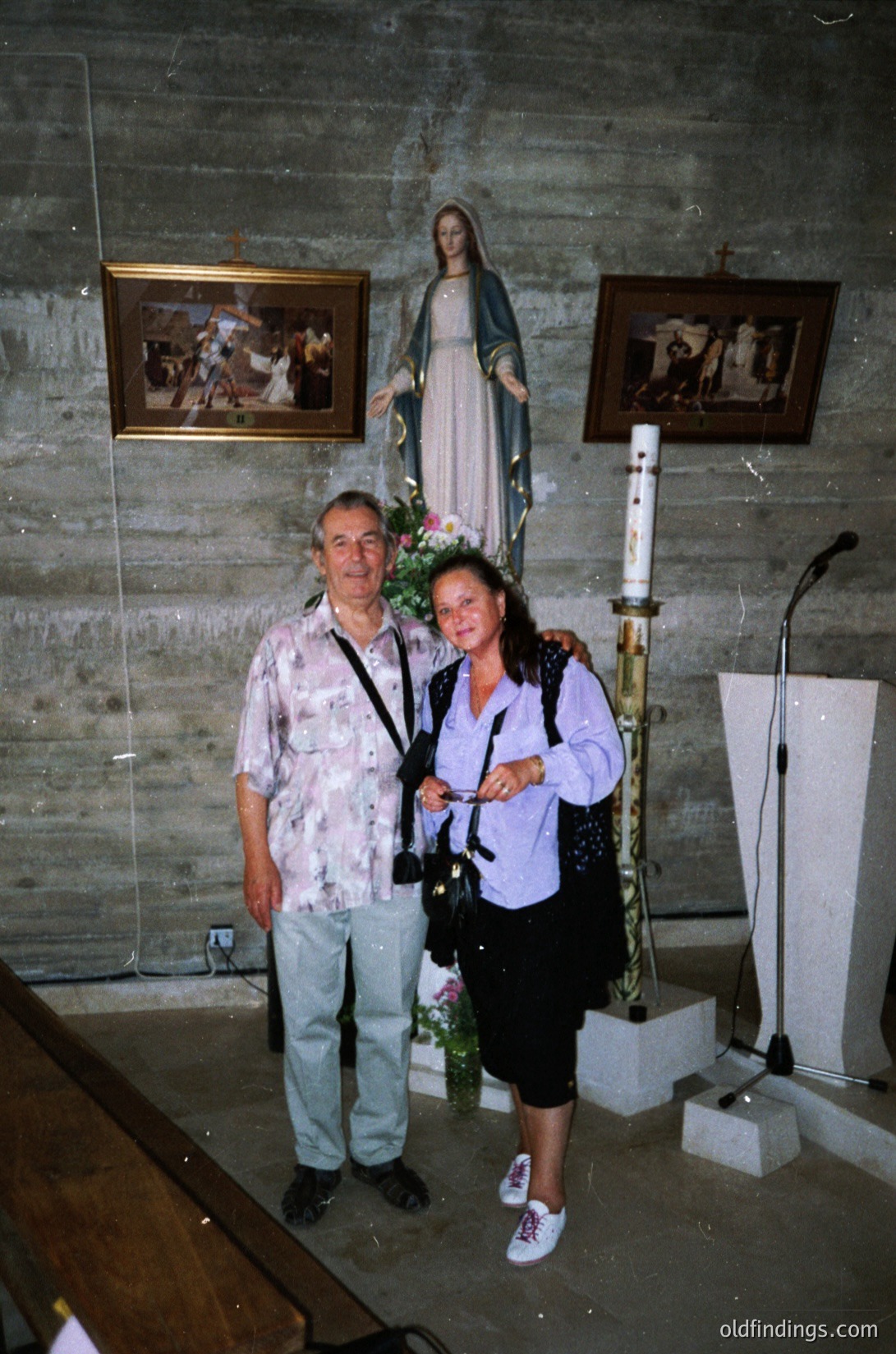 Two individuals pose indoors in a chapel-like setting, likely Eastern European (1990s). The man wears a floral shirt and jeans; the woman, a purple top with a camera strap. A statue of the Virgin Mary (central) and religious paintings flank them. Concrete walls and a lectern suggest a modest, utilitarian design.