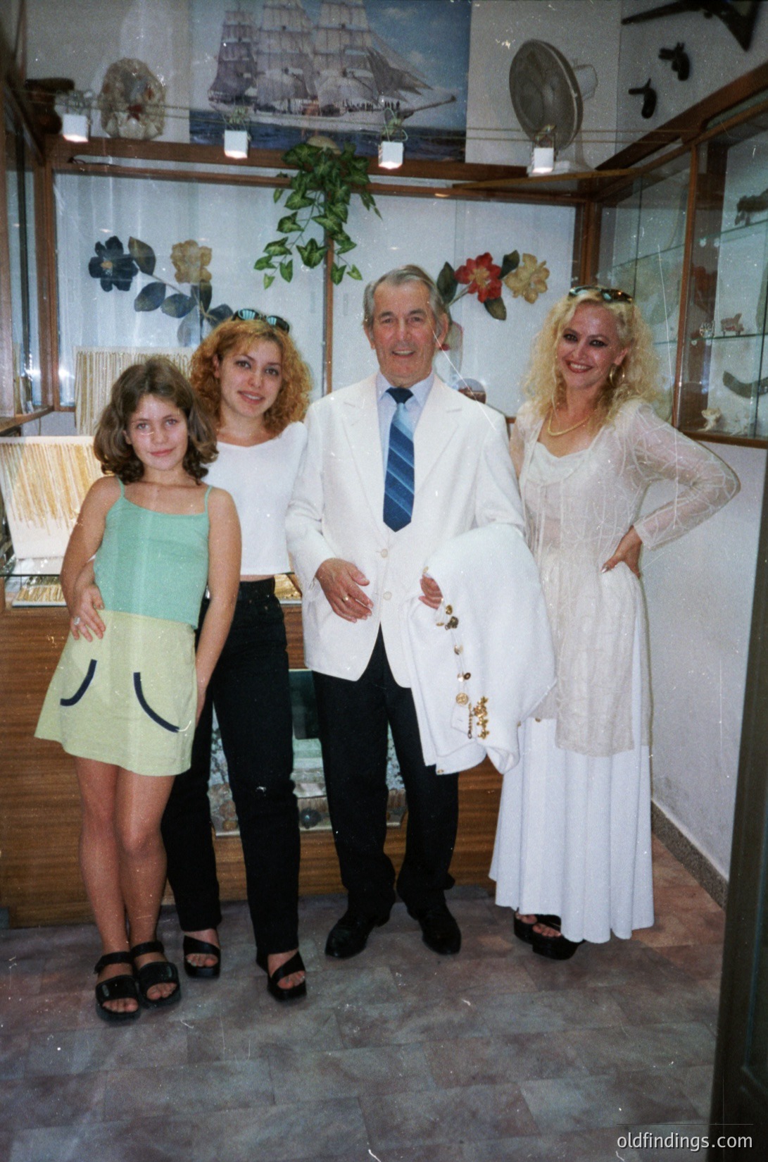 Family portrait in a vintage shop setting, likely 1980s–1990s. Four individuals pose: a man in a white suit with a striped tie, flanked by three women in casual-to-formal attire (green sleeveless dress, floral top, and white lace gown). Displayed behind them: maritime artifacts, framed paintings, and potted plants. Decor suggests a coastal or nautical theme.