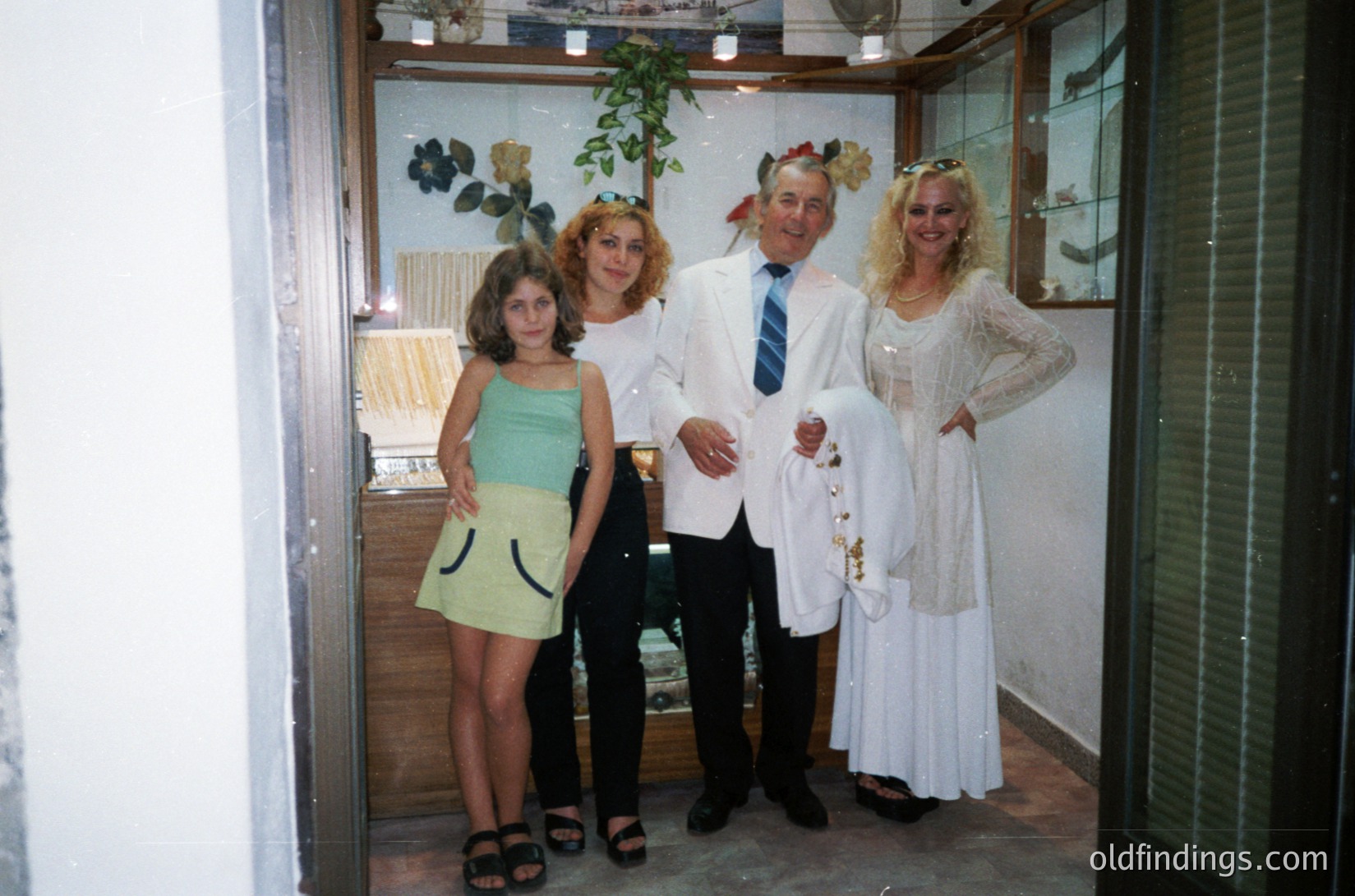 Family portrait in formal attire, likely a wedding or celebration, featuring four individuals: a young girl in a green strapless dress, a woman in a white blouse, a man in a white suit with a bow tie, and a woman in a white lace gown. Indoor setting with wooden paneling, potted plants, and decorative wall art. Style suggests 1970s–1980s Western/European influence.
