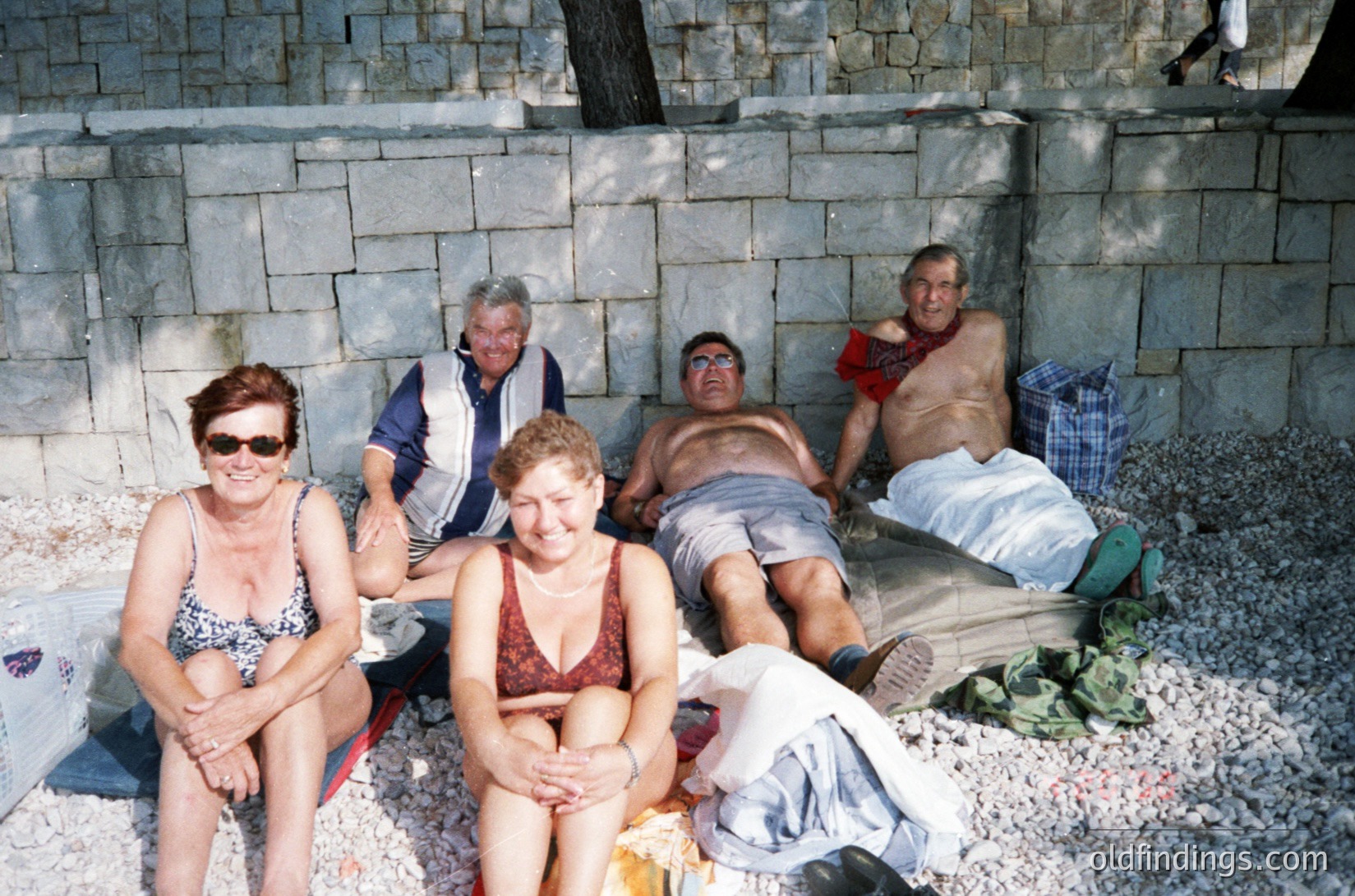 Five adults pose casually on a pebble beach beside a concrete wall, likely a seaside resort. The woman in the foreground wears a floral bikini, while others are in swimwear or shorts. One man holds a drink, suggesting a relaxed, social gathering. The concrete wall and pebble texture indicate a Mediterranean or Eastern European coastal setting, possibly the 1980s–1990s.