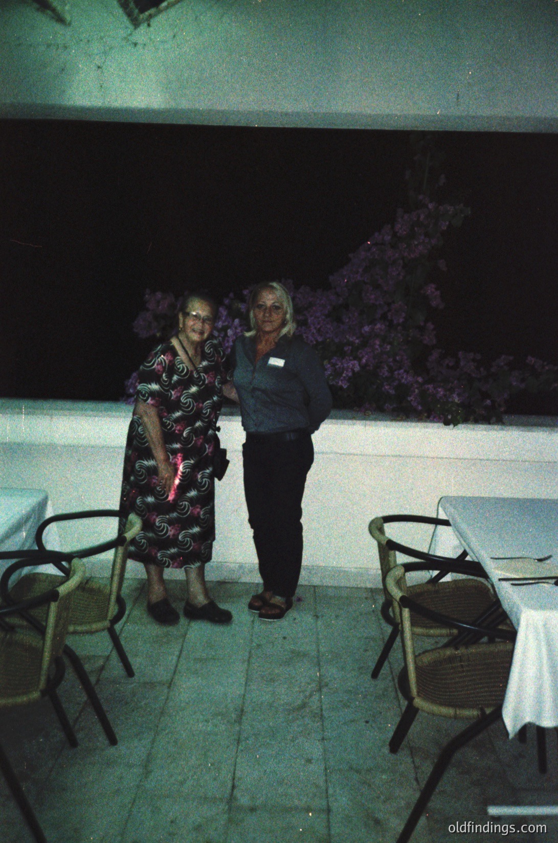 Two women pose outdoors at night under artificial lighting, likely a resort or hotel patio. The elder woman wears a patterned dress with a cane, while the younger woman in a dark polo stands beside her. Metal chairs and tables with white tablecloths surround them. Purple flowering vines decorate the background. [Mid-20th century resort evening scene with vintage attire and floral decor ]