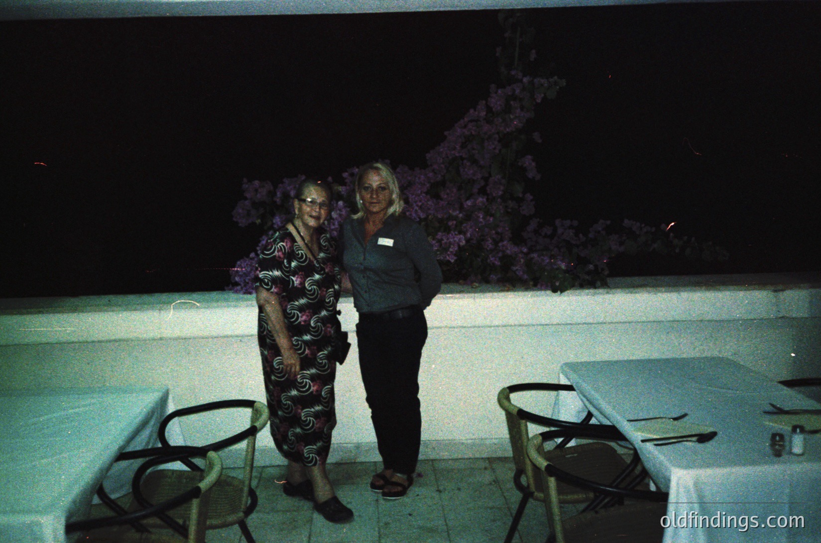 Two individuals pose outdoors at night under artificial lighting, likely at a resort or event venue. The woman on the left wears a patterned dress with floral motifs, while the man on the right wears a dark polo shirt with a name tag. A blooming purple flowering bush frames the background. Metal chairs and a table draped with a white cloth are visible, suggesting a dining or social area.