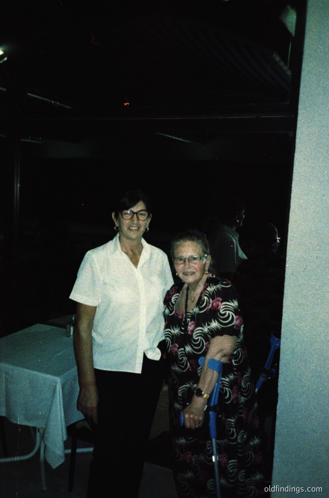 Two women pose indoors, likely in a 1970s-1980s setting. The woman on the left wears a short-sleeve blouse with a high collar, while the seated woman on the right dons a patterned, long-sleeve dress and holds a cane. A table with a white cloth is visible behind them, suggesting a dining or social area.