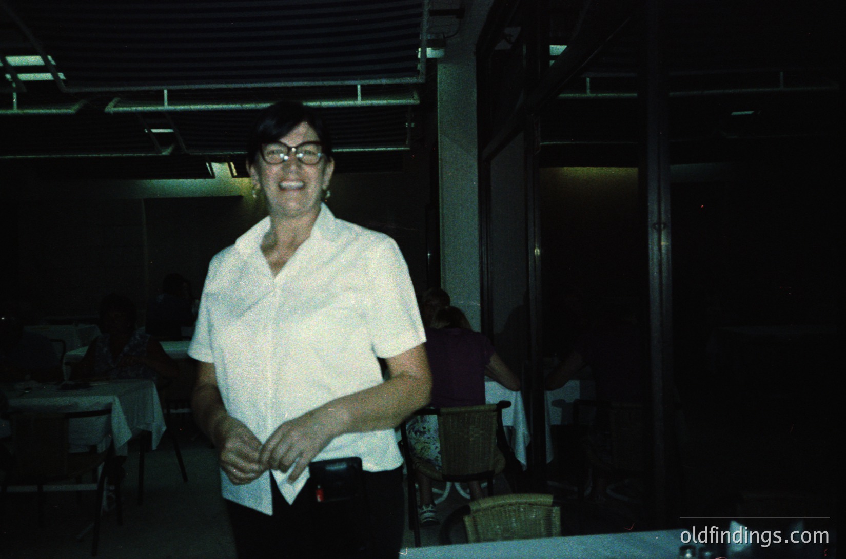 Mid-century indoor dining scene featuring a smiling woman in a white collared shirt and glasses, likely a waitress or hostess. Wooden tables, metal chairs, and a dimly lit, industrial-style ceiling with exposed beams. Possible 1960s–1970s restaurant or hotel lobby.