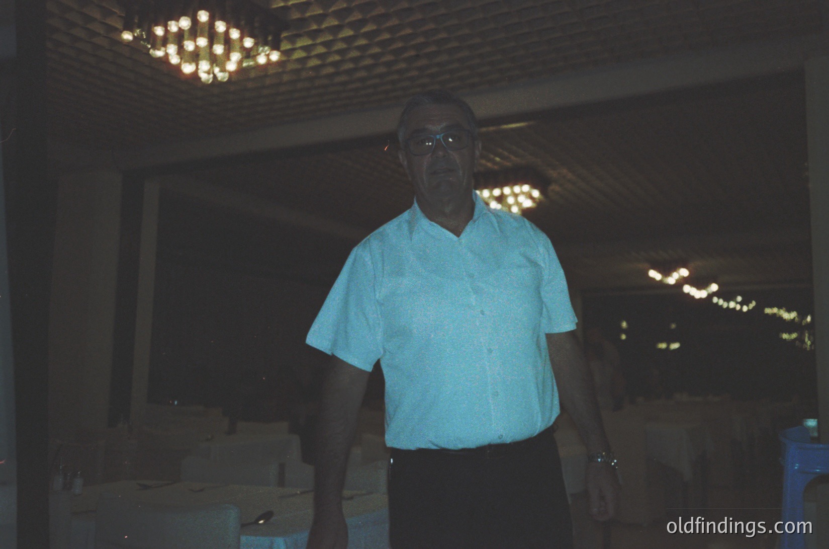 Mid-century man in a light blue button-down shirt poses indoors under a chandelier-lit ceiling. Stacked boxes and shelves suggest a storage or warehouse setting. Likely 1960s–1970s American or European style.