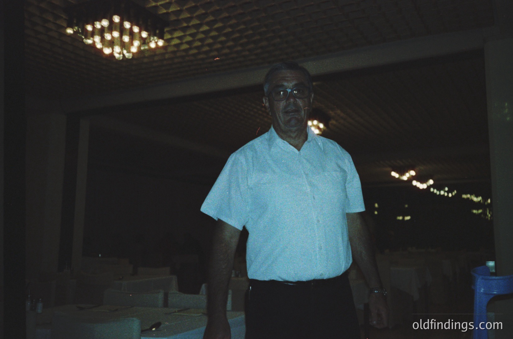 Mid-century interior portrait of an elderly man in a light-colored button-down shirt, standing in a dimly lit room with a chandelier and blurred outdoor lights. Style suggests 1960s–1970s era, possibly a café or lounge.