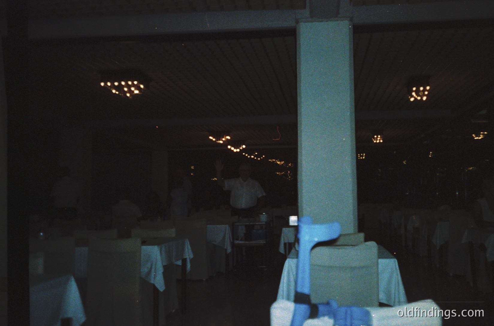 Vintage-style restaurant interior with high-backed chairs draped in blue tablecloths, string lights overhead, and a central pillar. Minimalist decor suggests 1970s-80s design. Empty tables imply evening setup or off-peak hours.
