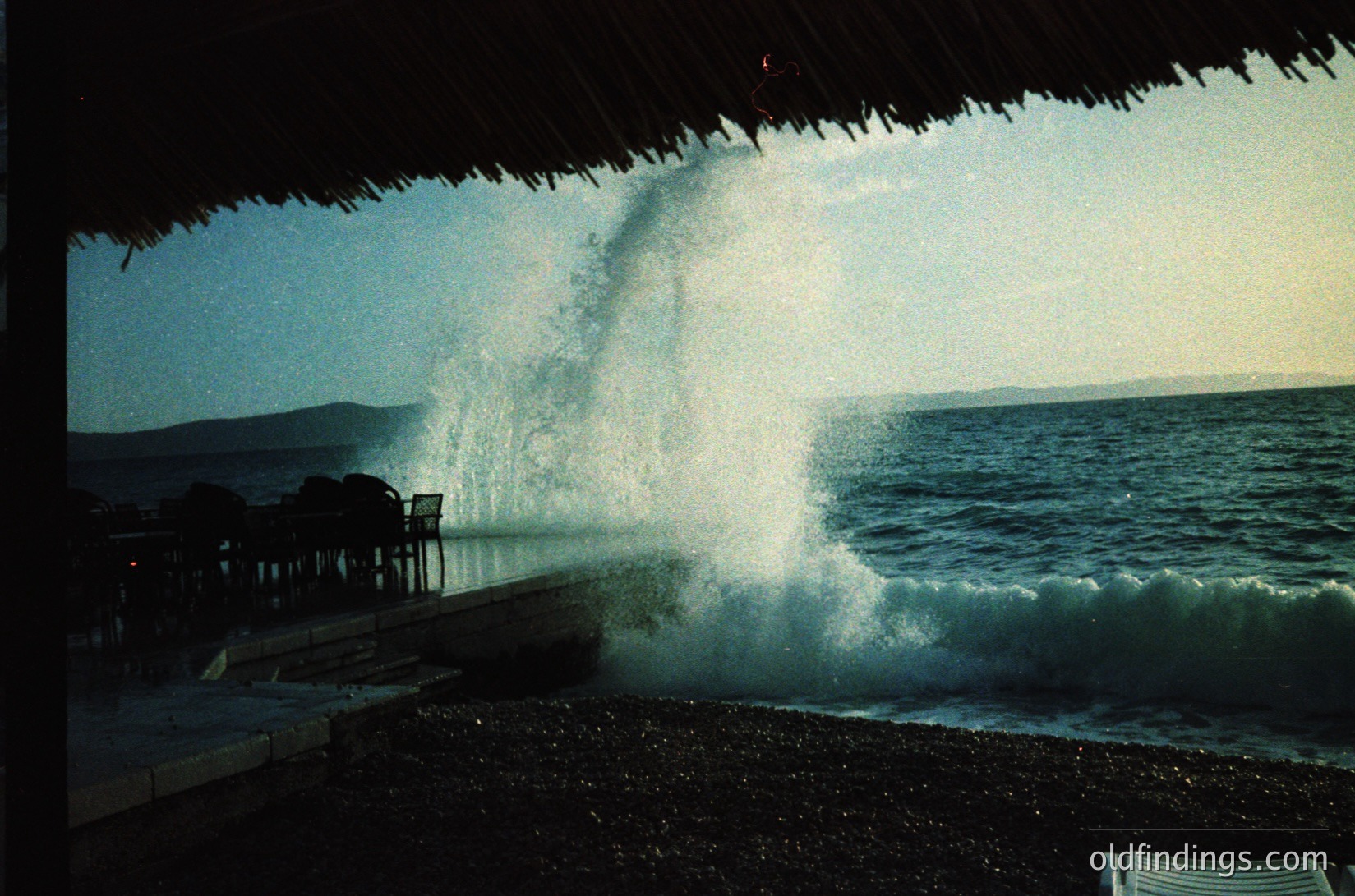 Seaside scene featuring a thatched-roof structure framing a powerful wave crashing onto a pebble beach. Ocean waves dominate the right, with distant landforms visible under a clear sky. Likely a coastal resort or beachside café.