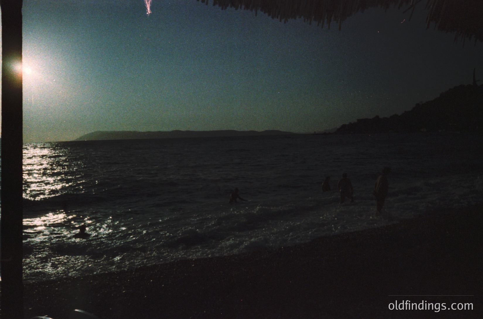 Silhouetted figures wade in shallow waves at golden-hour seaside. Low-angle sunlight reflects off water, illuminating foam. Coastal silhouette features rocky terrain. Likely mid-20th century beach scene.