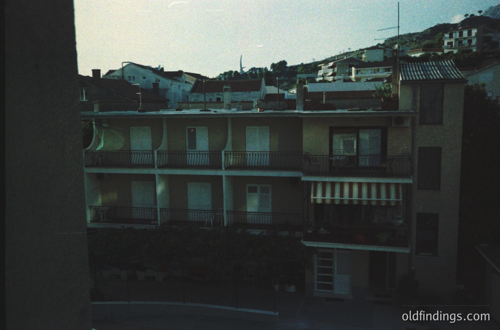 Mid-century Mediterranean urban rooftops with concrete balconies and tiled roofs, viewed from an interior window. Residential buildings cluster on a hilly terrain, suggesting coastal or seaside proximity. Architectural style hints at 1960s–1970s construction.