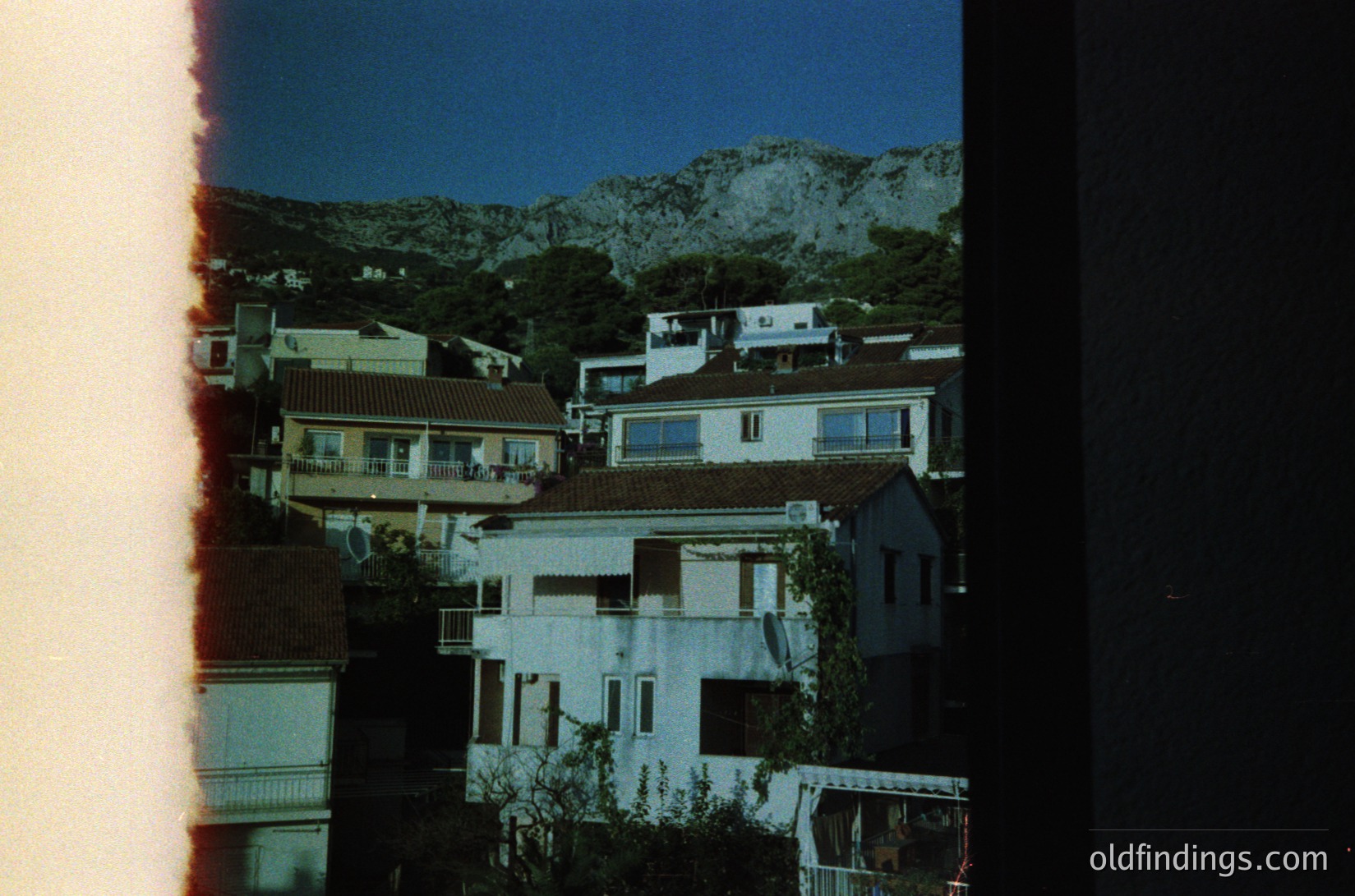 Vintage film scan of Mediterranean hillside village at dusk, featuring clustered whitewashed buildings with terracotta roofs. Rugged limestone mountains frame the scene, suggesting a coastal or inland Mediterranean climate. Architectural style hints at 20th-century local construction.