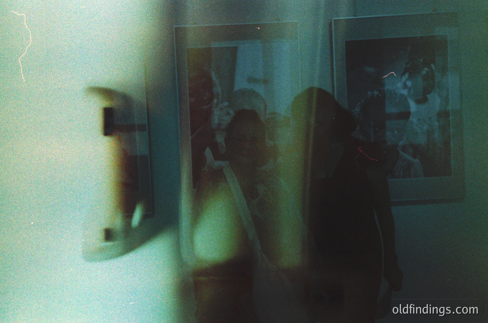 Vintage Polaroid-style photo through a frosted glass door, revealing a blurred indoor scene with a woman in a patterned dress and a child. Reflections of framed photos on the wall show a group of people. Likely mid-20th century domestic setting.