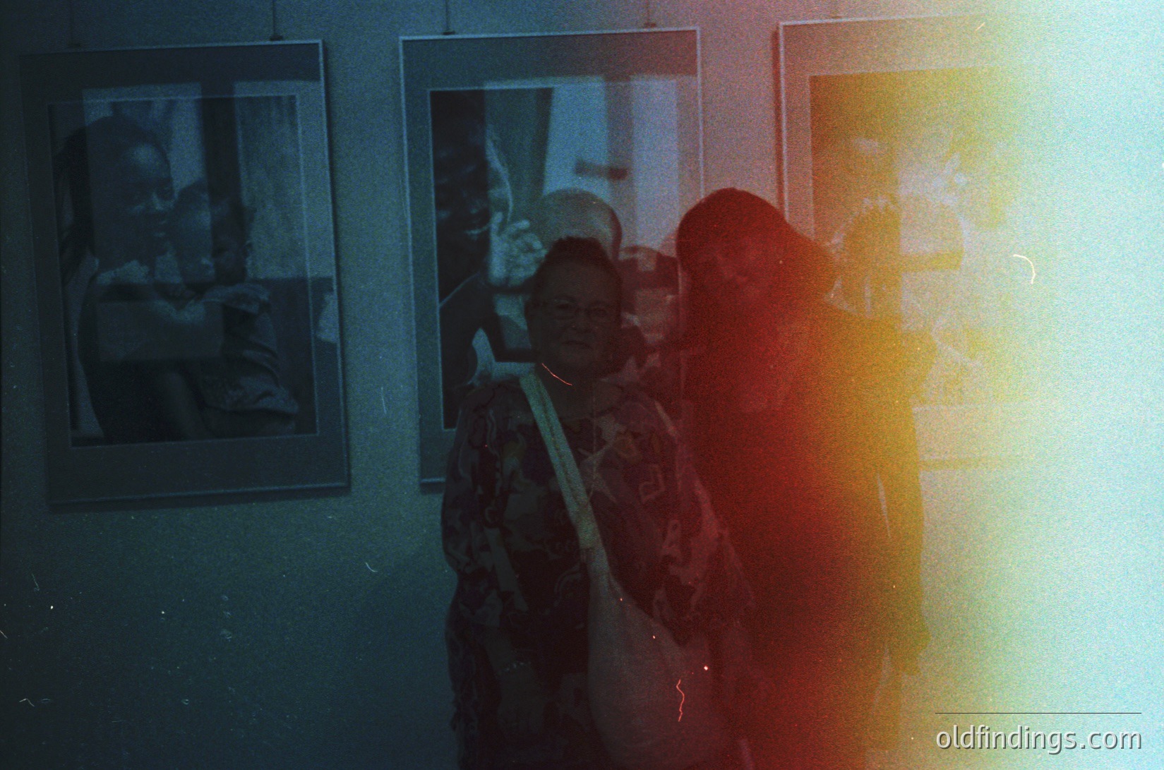 Vintage-style photo of two figures in a dimly lit gallery, framed by warm sepia tones. One wears a patterned jacket; both stand close to framed artwork depicting urban scenes. Reflections and lens flare enhance nostalgic atmosphere. Likely 1970s–1980s, urban setting.