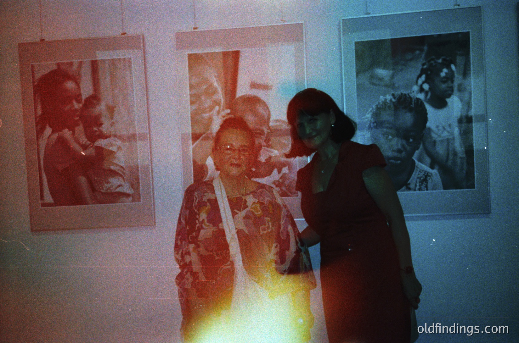 Vintage exhibition featuring framed black-and-white portraits on a dimly lit wall, likely from the 1960s–1970s. Central figure in patterned blouse poses with a younger woman in dark clothing. Warm light flare enhances nostalgic atmosphere. Ideal for historical research or vintage aesthetic projects.