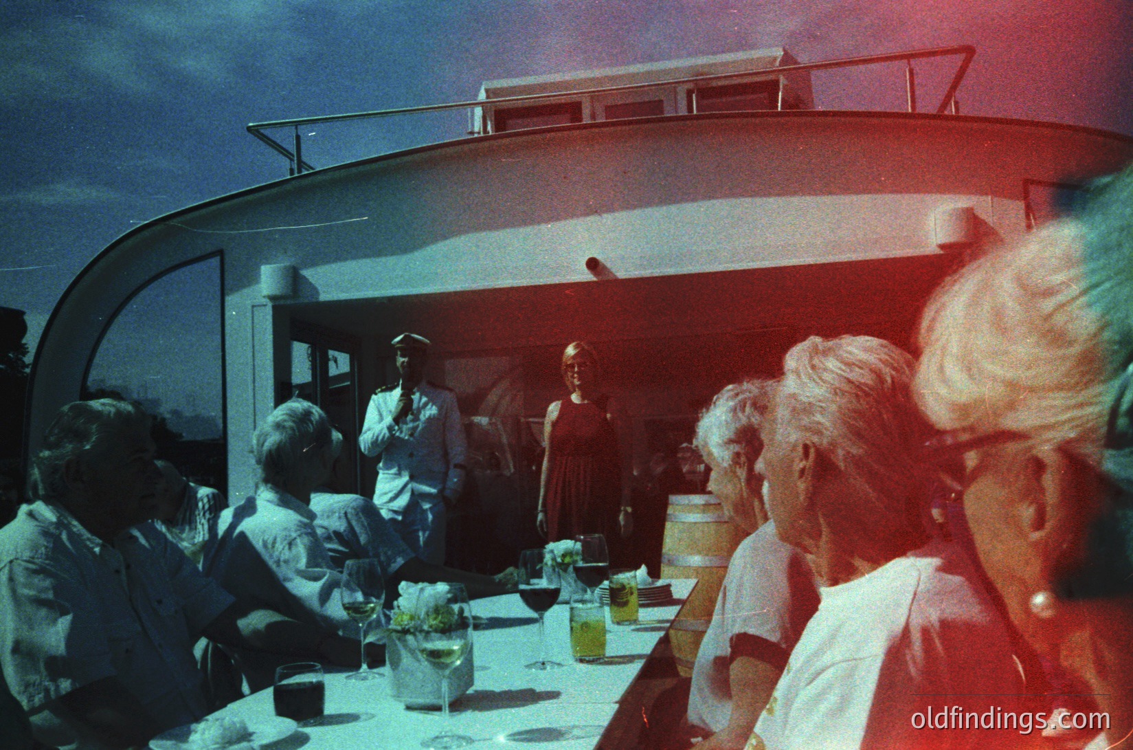 Vintage cruise ship dining hall with mid-century modern curved ceiling. Passengers in 1960s attire—men in suits, women in dresses—gather around a long table set with glasses, plates, and floral centerpieces. A waitstaff member in uniform serves food. Warm, nostalgic lighting enhances the retro ambiance.