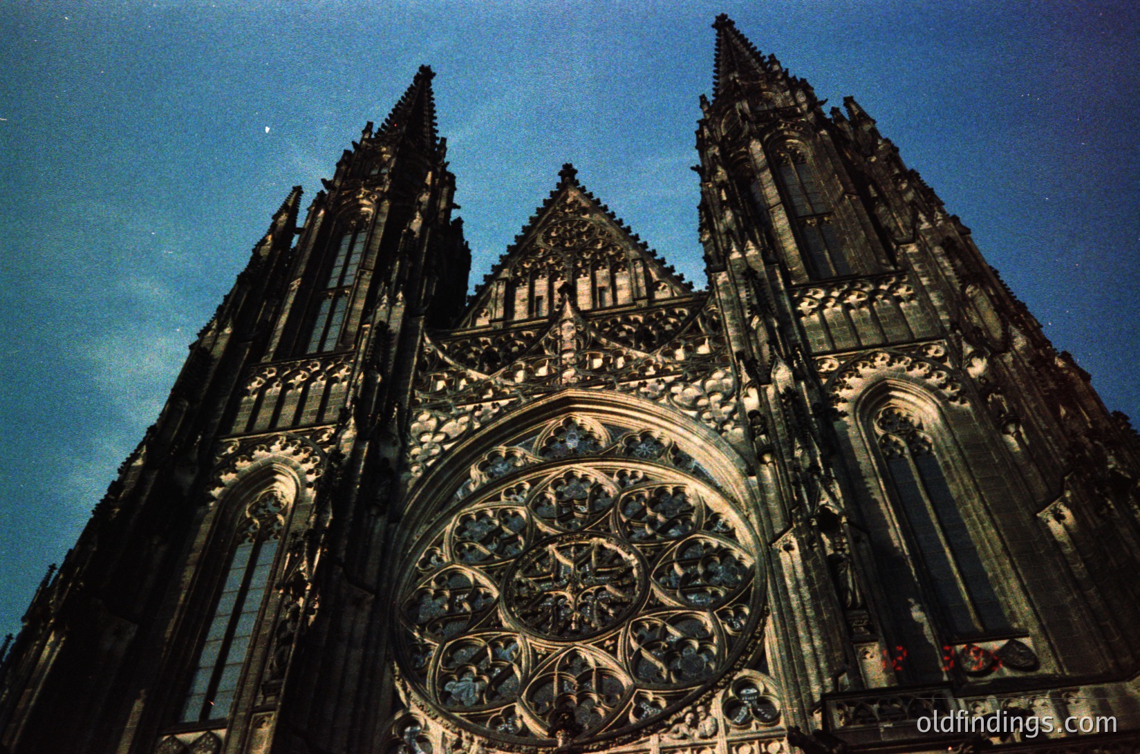 Gothic cathedral façade featuring intricate stonework, twin spires, and a grand rose window with tracery. Likely European, 13th–16th century. Symmetrical buttresses and pointed arches dominate the design.