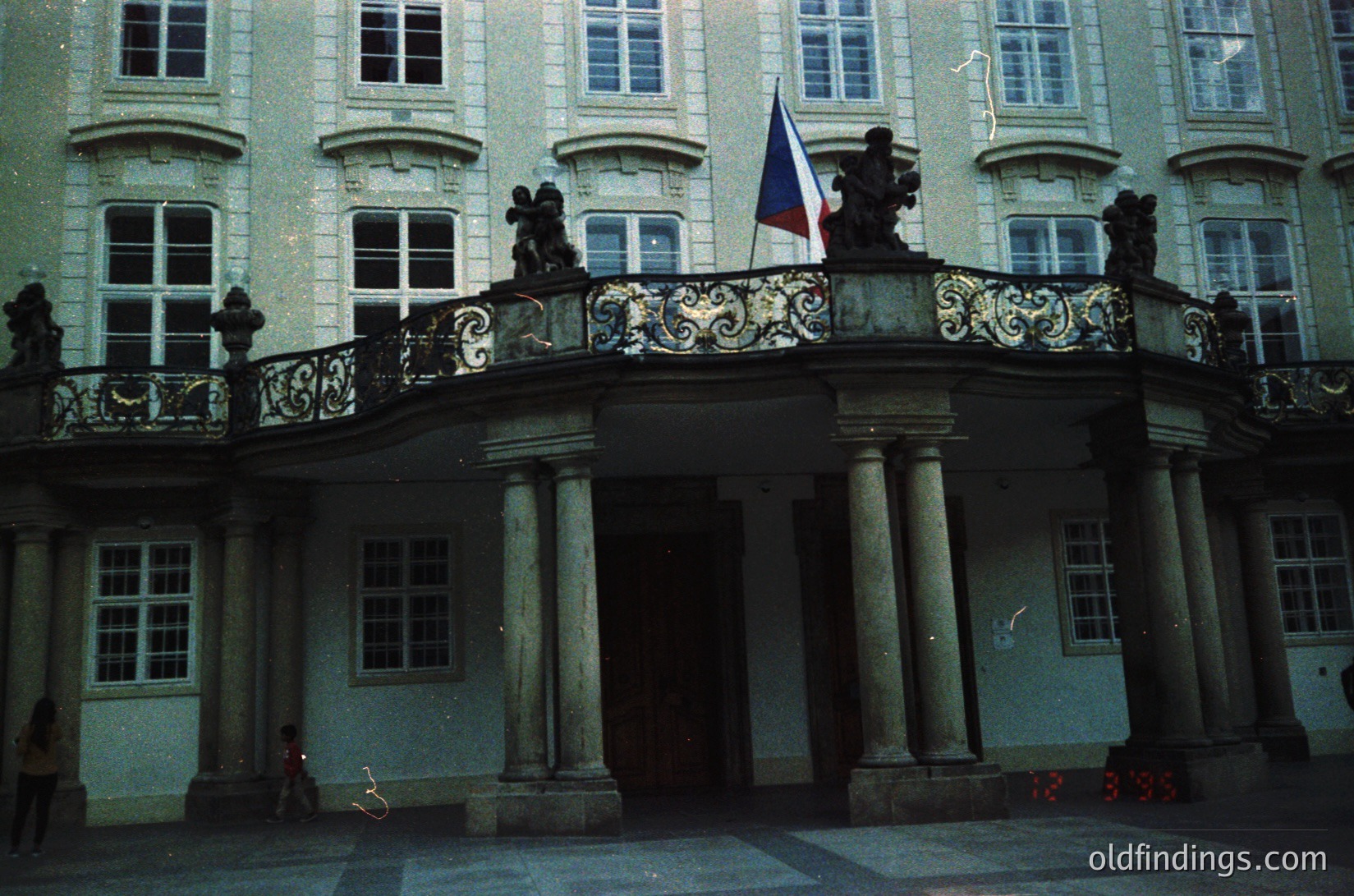 Neoclassical courtyard facade featuring ornate balustrade with sculpted urns, flanked by tall Corinthian columns. A flagpole displays a tricolor flag. Symmetrical windows with divided panes. Likely European institutional or governmental building, 19th–early 20th century.