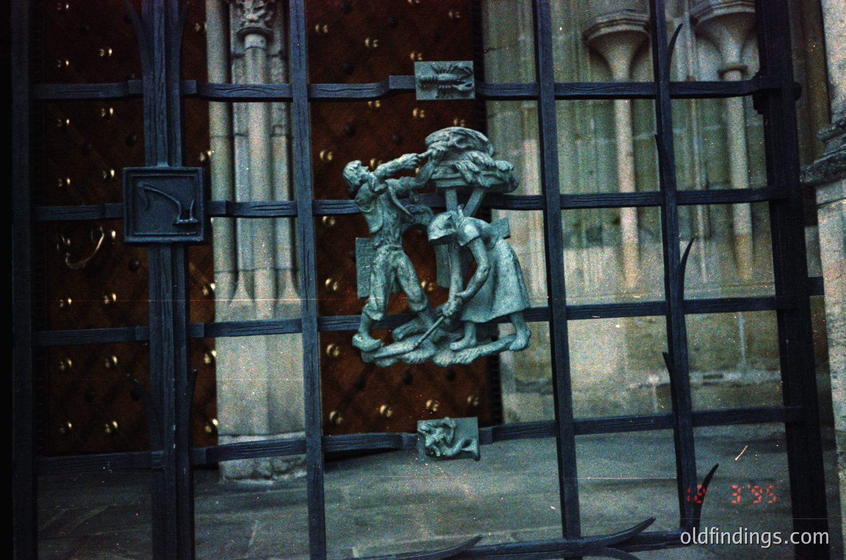 Relief sculpture of two figures in dynamic pose on ornate wrought-iron gate, likely 19th-century European. Figures appear to be engaged in labor or conflict, framed by Gothic-style columns. Stained glass and architectural details visible through gate.