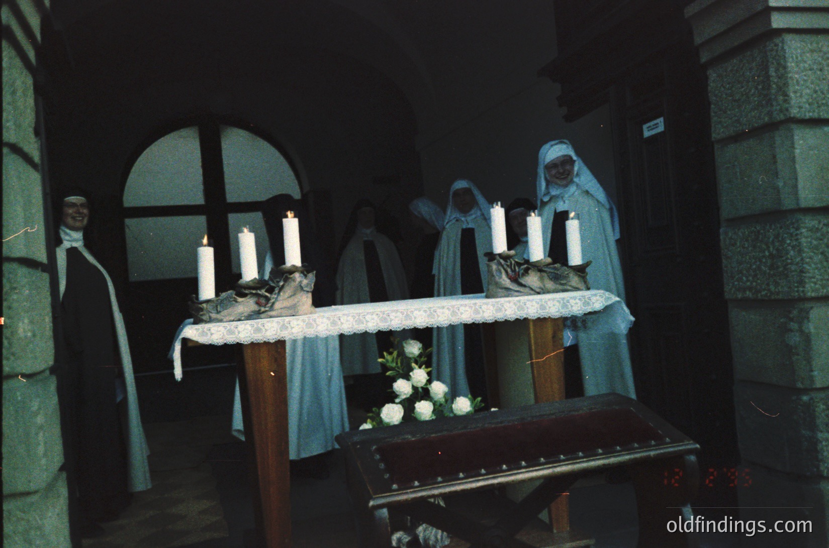 Religious procession altar featuring nuns in traditional habits, holding lit candles. Wooden lectern draped in white cloth with white flowers. Gothic-style stone archway and doorway in background. Likely Eastern Orthodox or Eastern Catholic liturgical setting.