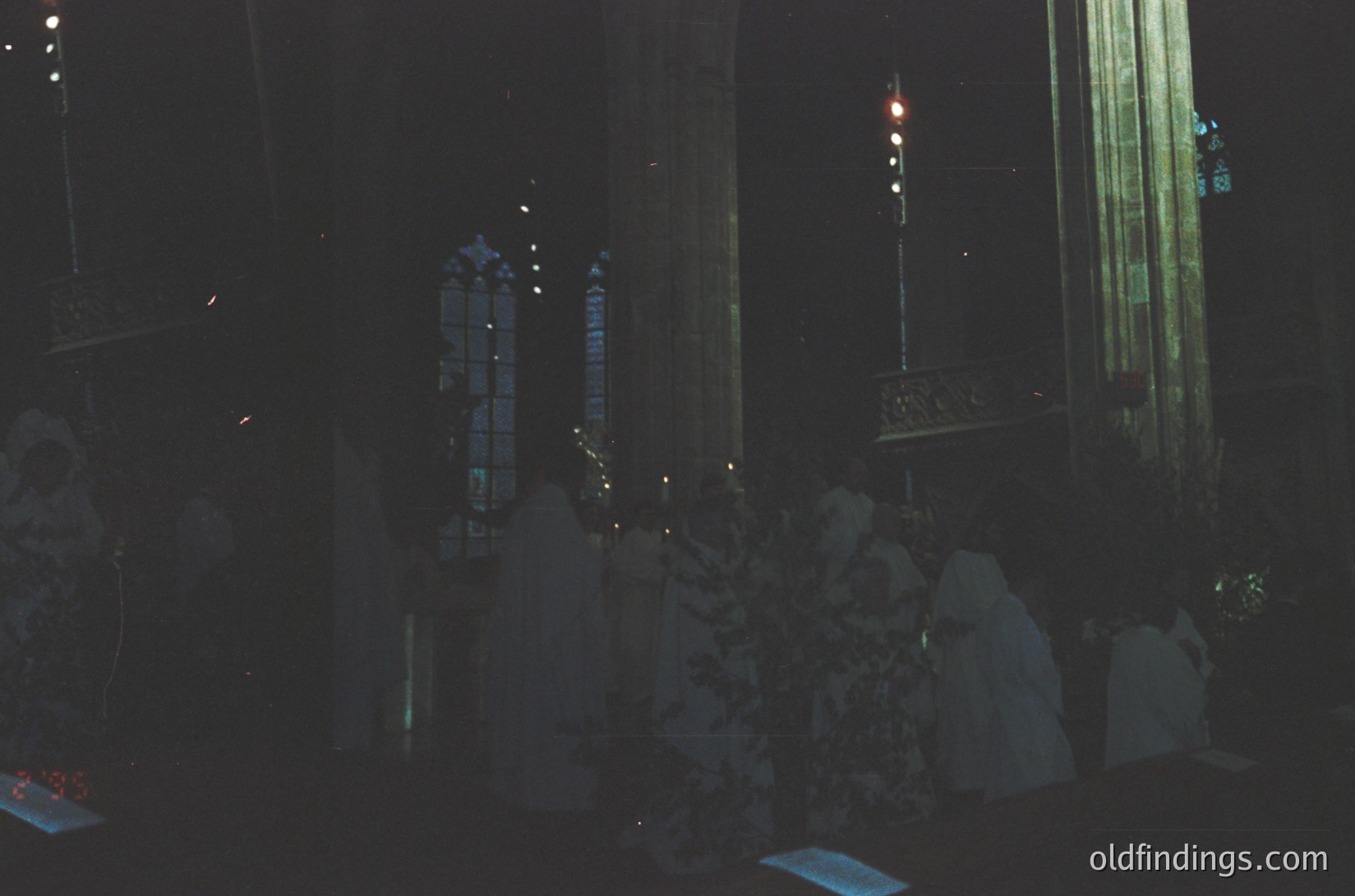 Nighttime interior of a grand, illuminated church with vertical stained-glass windows casting colored light. Ornate chandeliers and intricate architectural details highlight Gothic Revival style. Crowd of silhouetted figures in formal attire, likely attending a ceremony or concert.