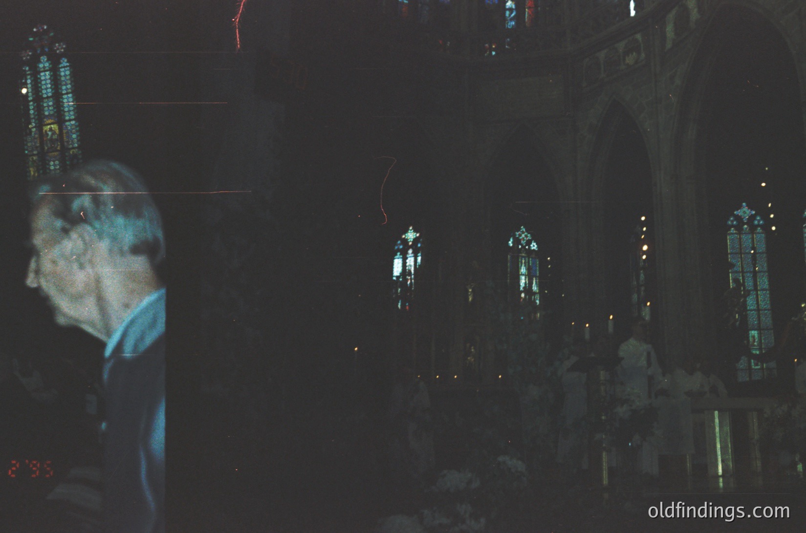 Blurred silhouette of a person in a blue shirt inside a dimly lit Gothic-style church, illuminated by stained-glass windows casting colored light. Long exposure captures light trails from moving objects. Architectural details include pointed arches and ornate stonework. Likely 20th-century European church interior.