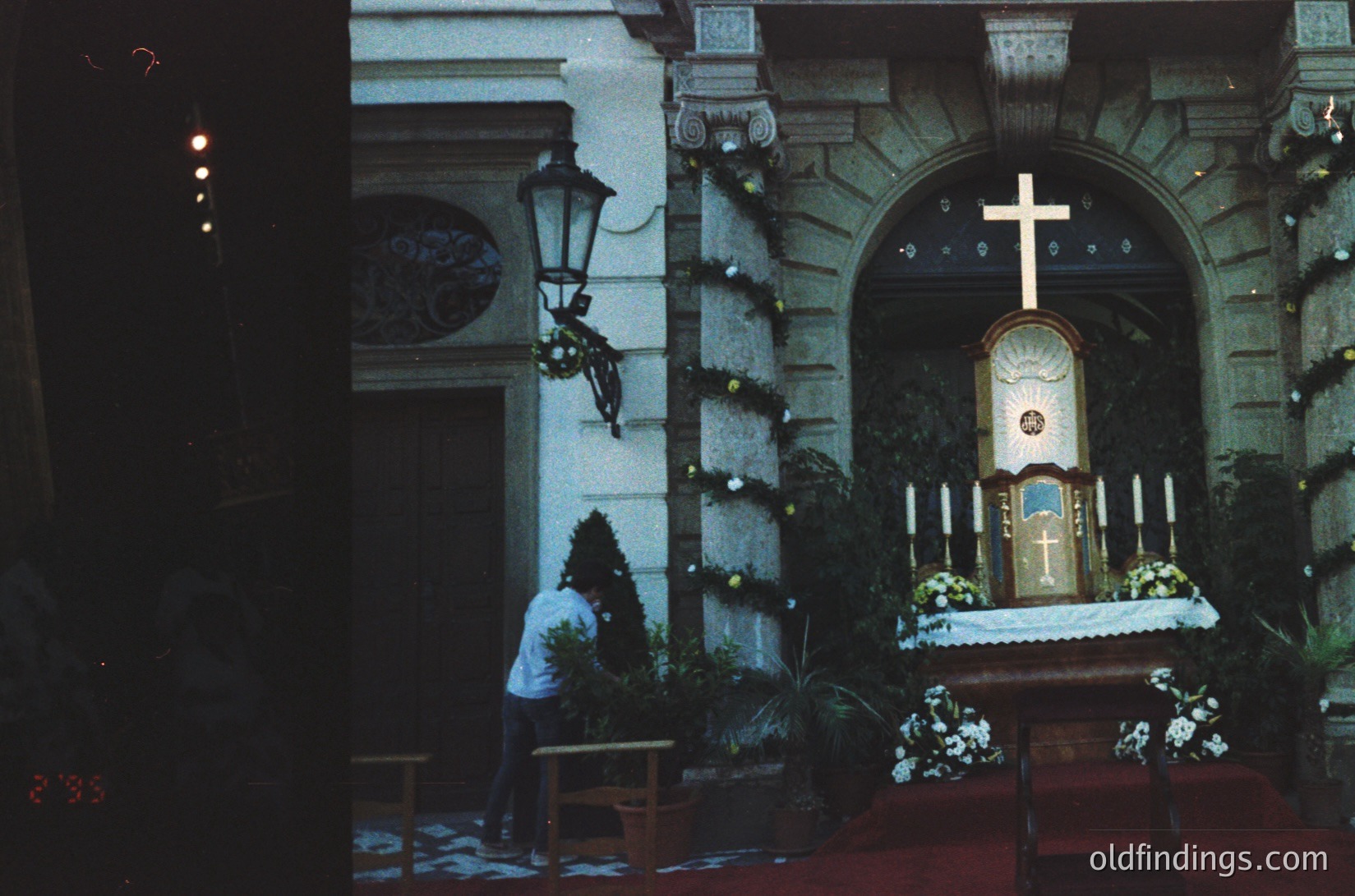 Vintage black-and-white church interior with ornate altar adorned with floral arrangements and a prominent cross. A priest in traditional vestments kneels in prayer at a side bench. Classic lantern and wrought-iron details frame the scene. Likely Eastern European, mid-20th century.