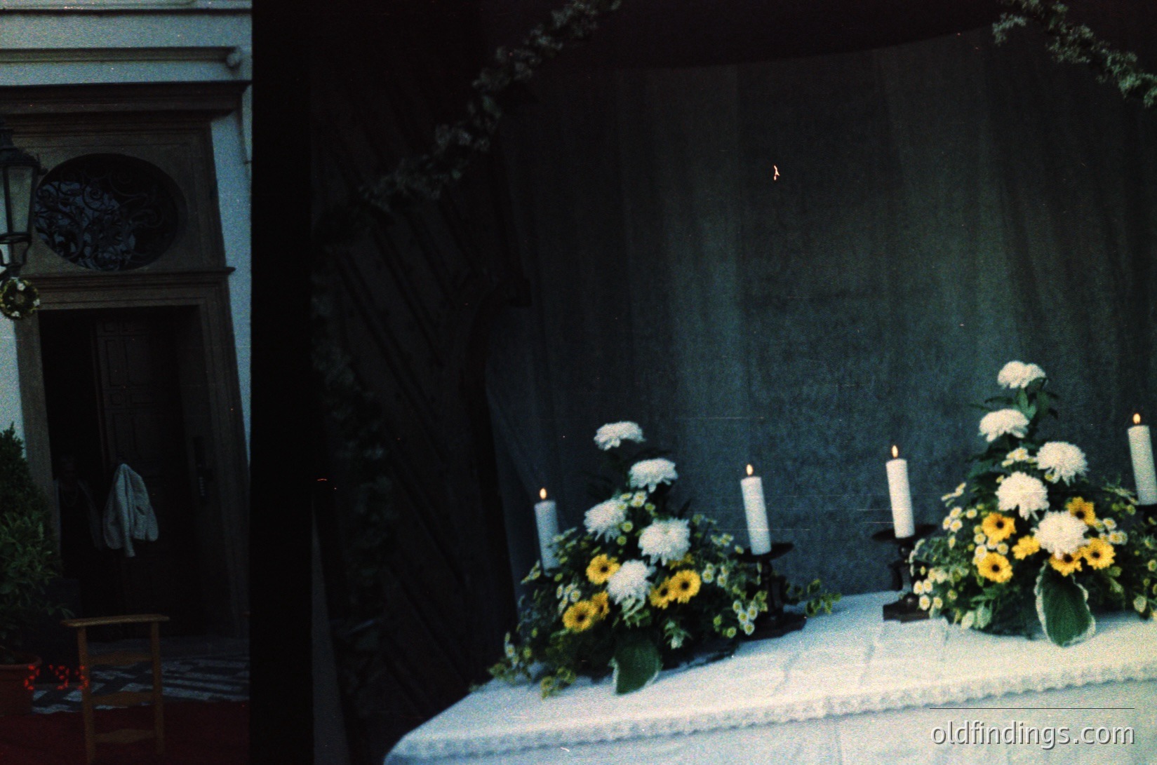 Symmetrical floral arrangements with white roses, yellow sunflowers, and greenery flank a draped altar table, lit by three lit candles. Ornate doorway with arched window and vintage lantern in a dimly lit interior, likely a church or chapel.