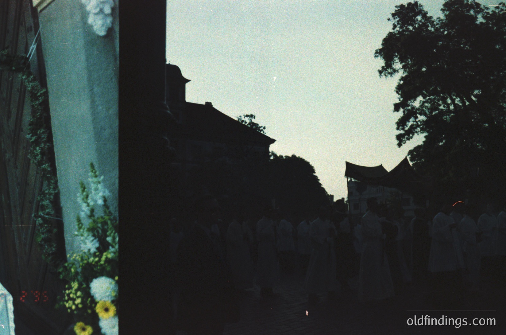 Vintage Polaroid-style shot of a dimly lit alleyway framed by a white picket fence and lush greenery. Sunlight filters through leaves, casting soft shadows on brick walls. Distinctive 1970s aesthetic with warm sepia tones. Urban greenery meets residential architecture.