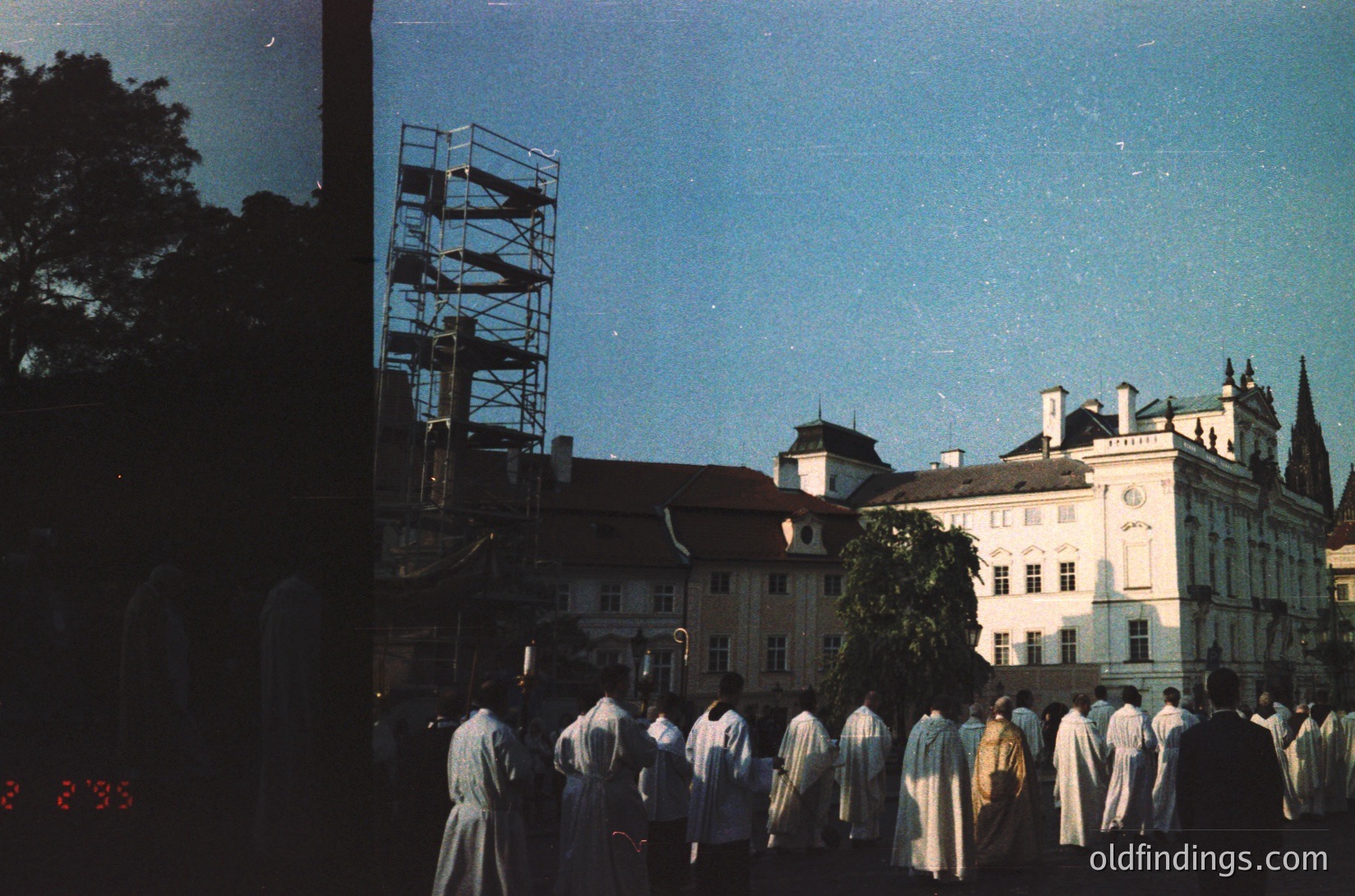 Vintage sepia-toned photo of a formal procession in a European courtyard, likely 1950s–1960s. Uniformed men in long coats march past a grand Baroque-style building under scaffolding. Clear blue sky and trees frame the scene.