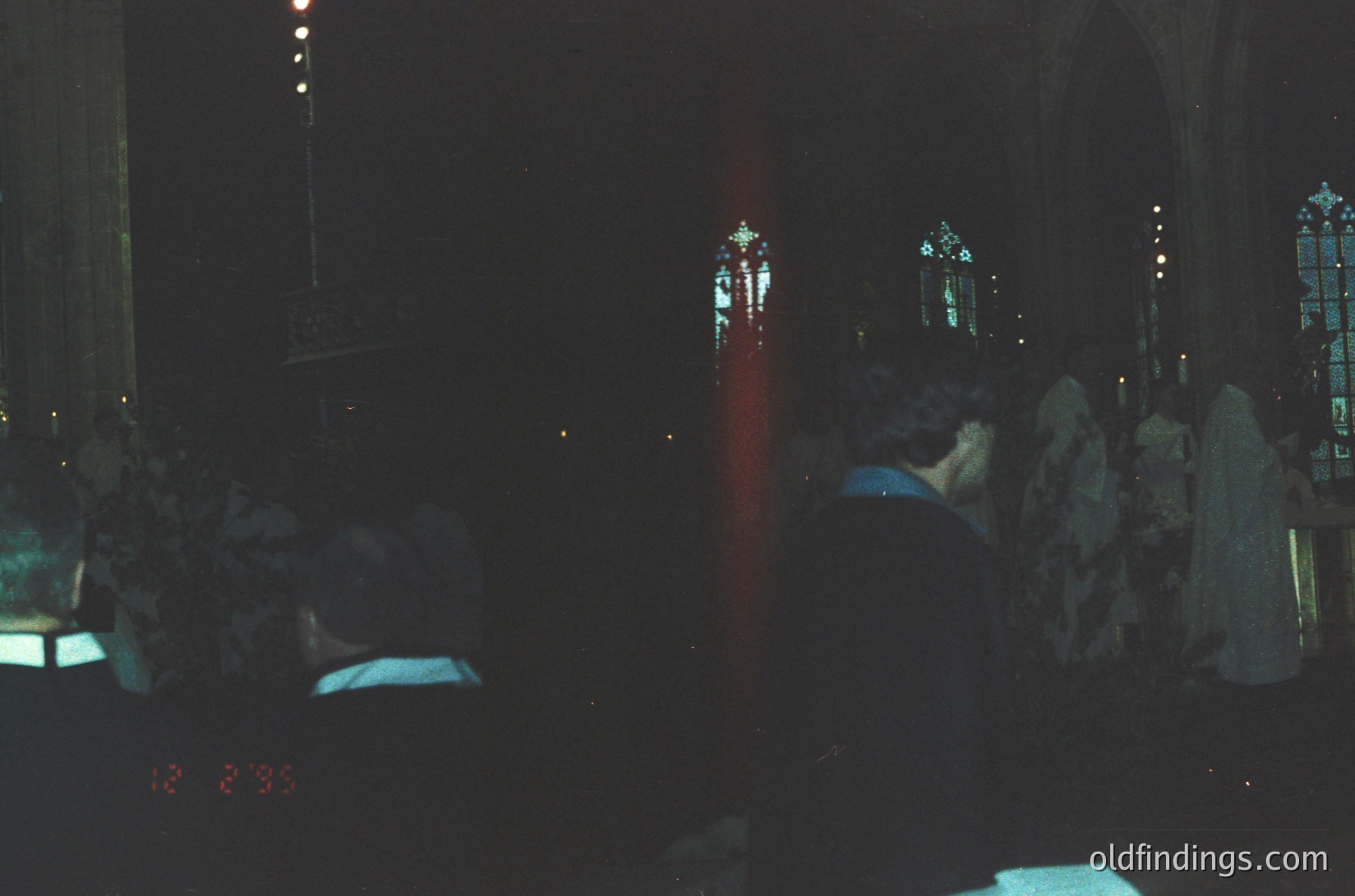 Gothic cathedral interior at night, illuminated by stained-glass windows casting colored light. Crowd seated in pews, facing altar with prominent crucifix. Low-angle shot highlights verticality of architecture. Likely European, 19th–20th century.