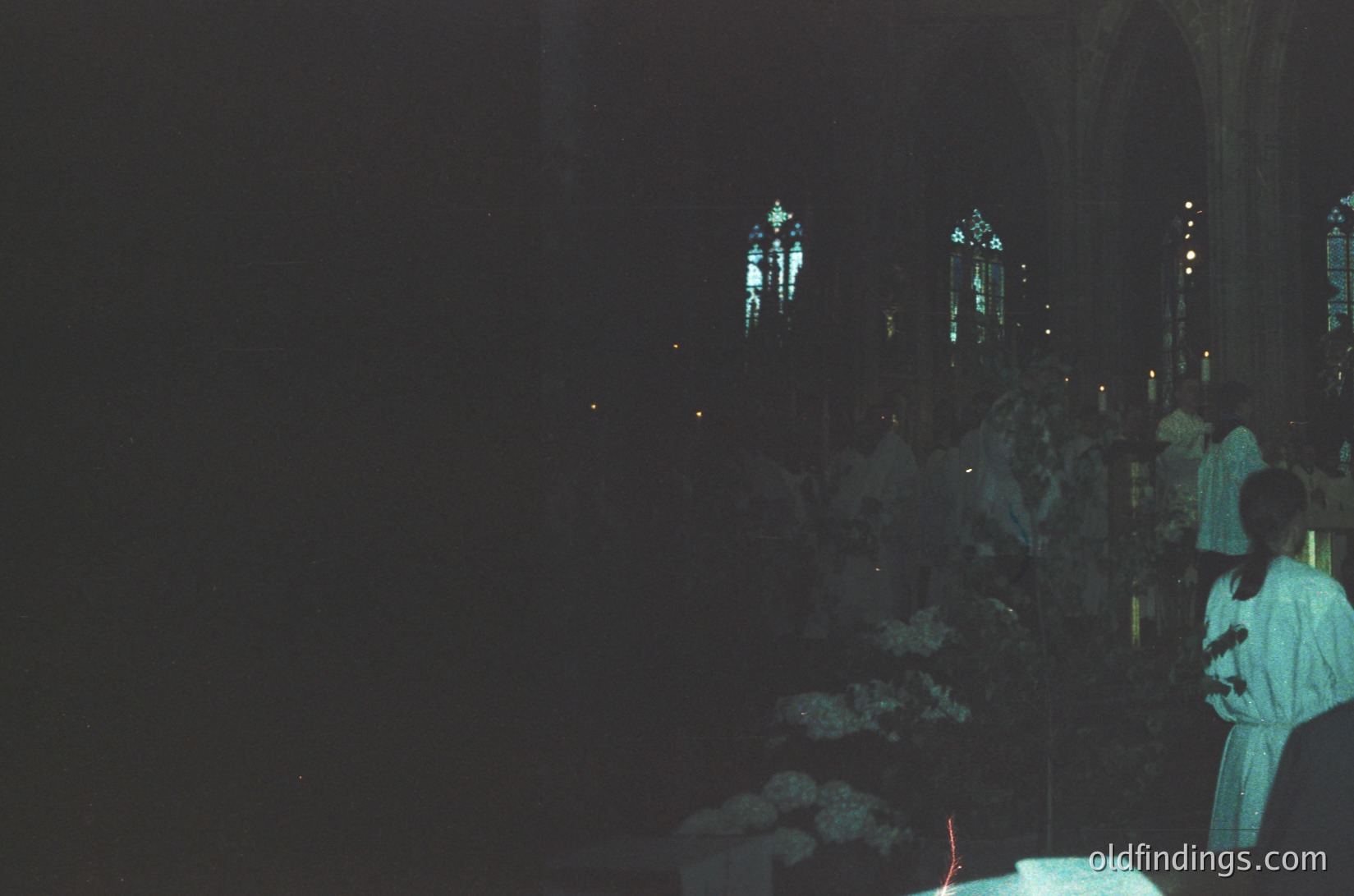 Gothic cathedral interior bathed in dim candlelight, showcasing stained-glass windows depicting religious figures. A priest in liturgical vestments conducts a service, with floral arrangements on the altar. Likely 19th–early 20th century European architecture.