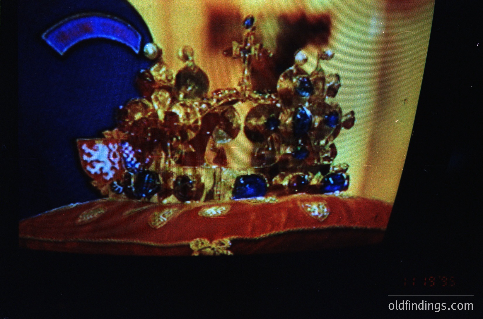 Elaborate crown adorned with gold filigree, sapphires, and emeralds, displayed under dramatic lighting. Intricate heraldic designs visible on the base. Likely European royal regalia, 18th–19th century.