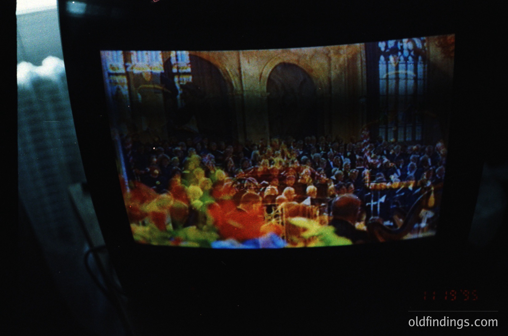 Vintage film strip projection of a packed concert hall, featuring a large orchestra and audience in a grand, arched interior. Warm stage lighting contrasts with dimmed seating. Likely 1950s–1970s, Western European venue.